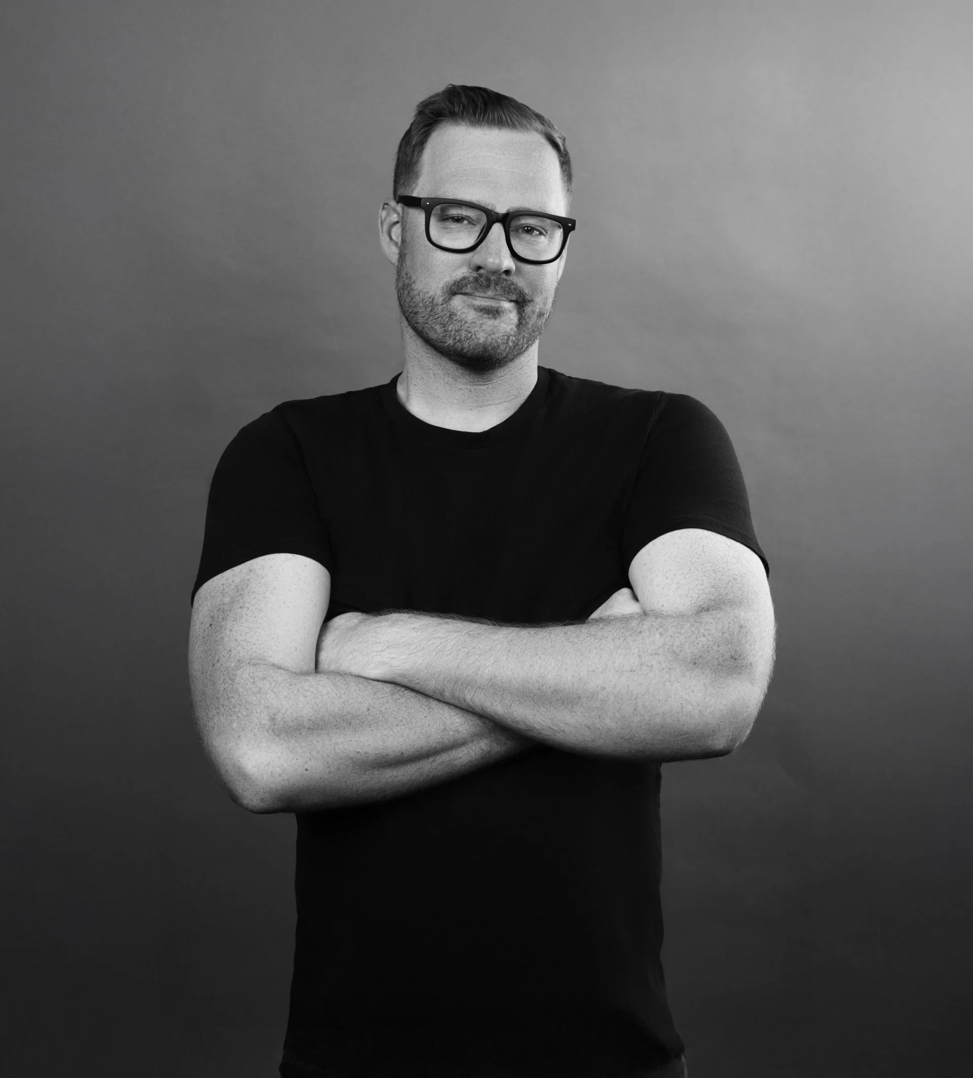 Black and white portrait of a man with glasses, short hair, and a beard, standing with arms crossed against a plain background.