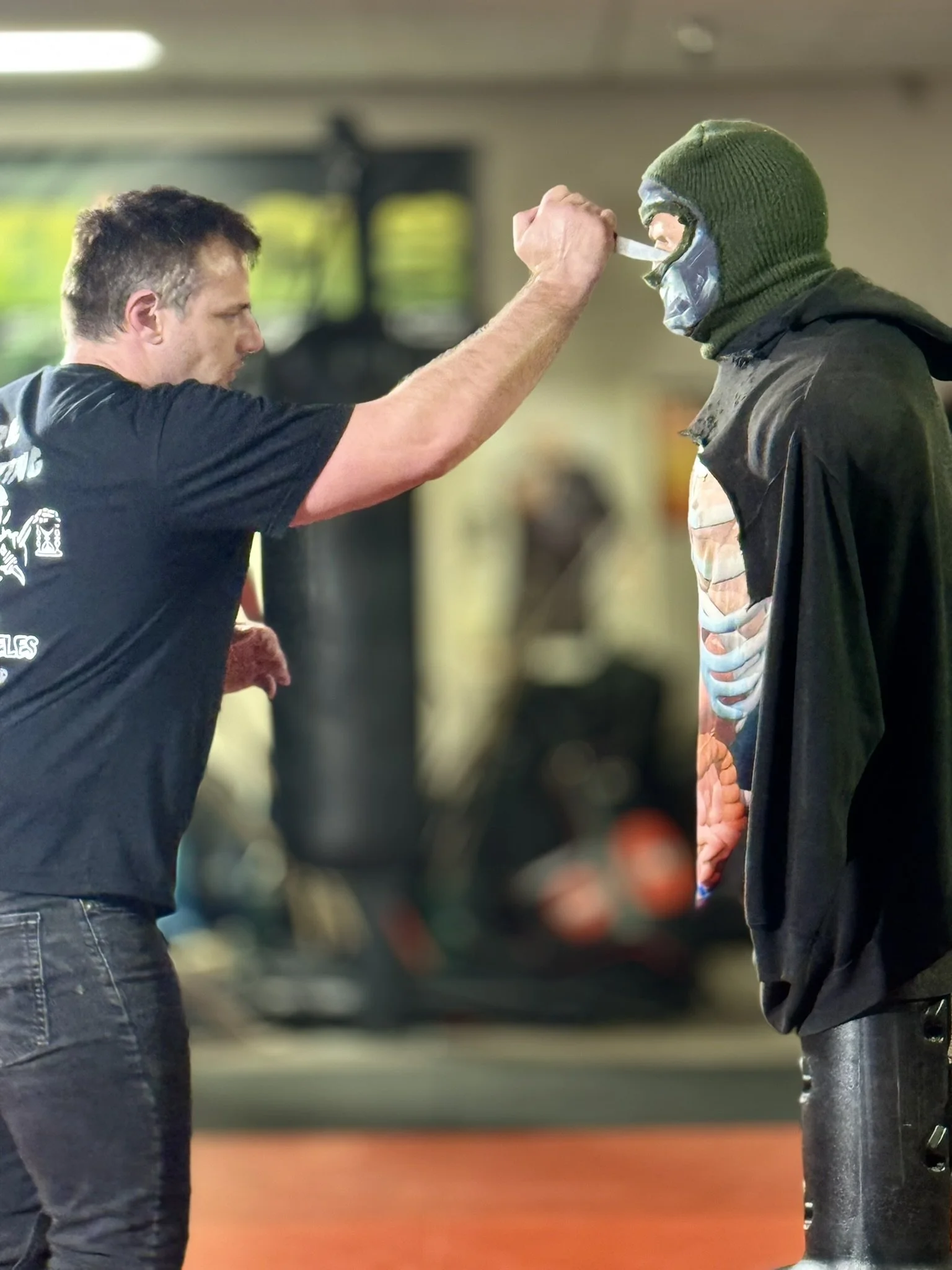 A person with short dark hair and a black t-shirt trains with a martial arts dummy wearing a green ski mask. The person is holding a knife and appears to be practicing a combat move in an indoor training facility.