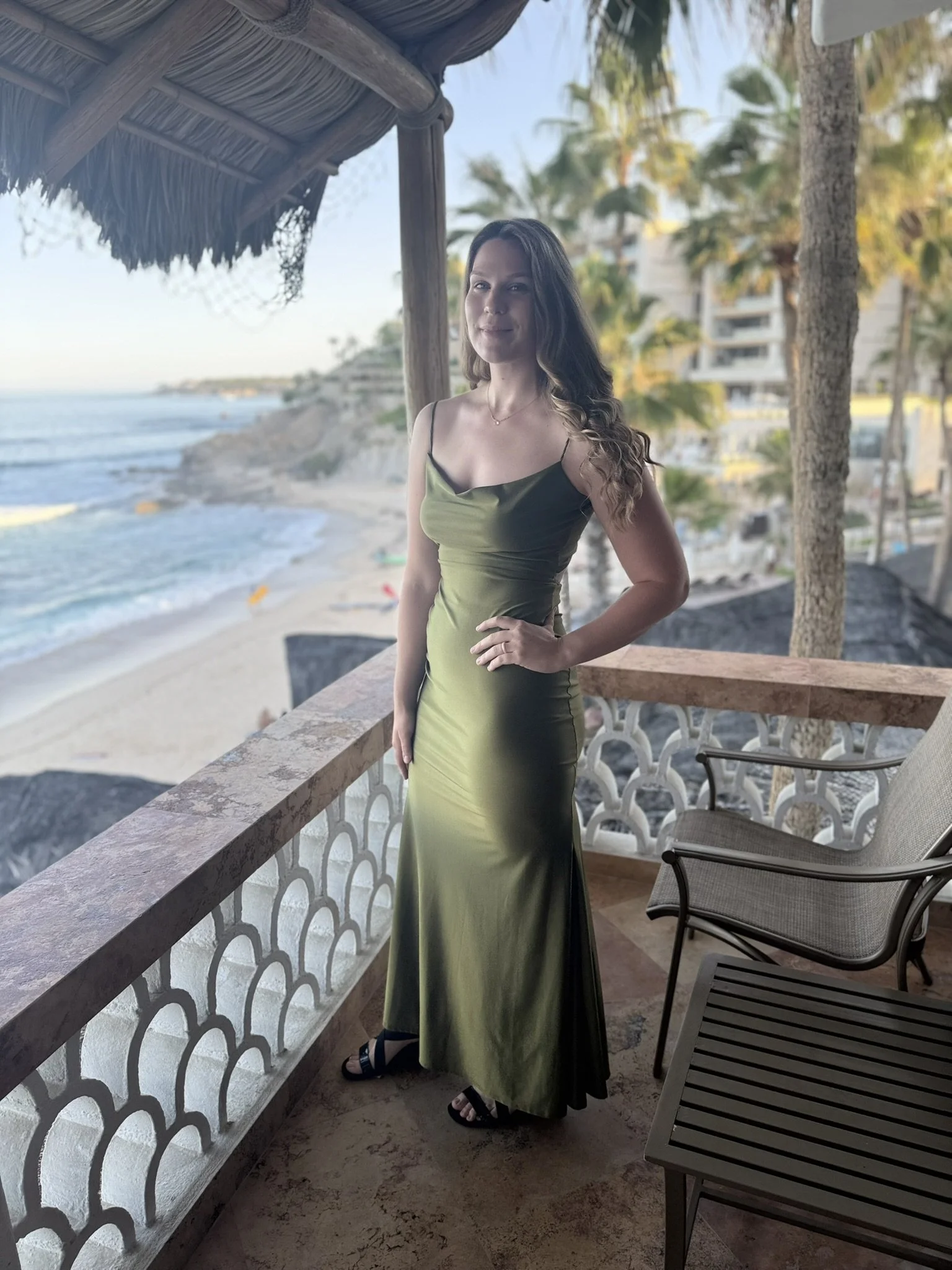 A woman with long dark hair standing on a balcony overlooking a beach with palm trees, ocean, and a clear sky, dressed in a green dress and black sandals.
