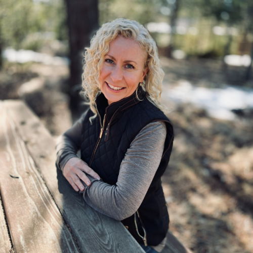 A woman with curly blonde hair smiling outdoors, wearing a black vest and gray long sleeve shirt, leaning on a wooden railing in a forested area