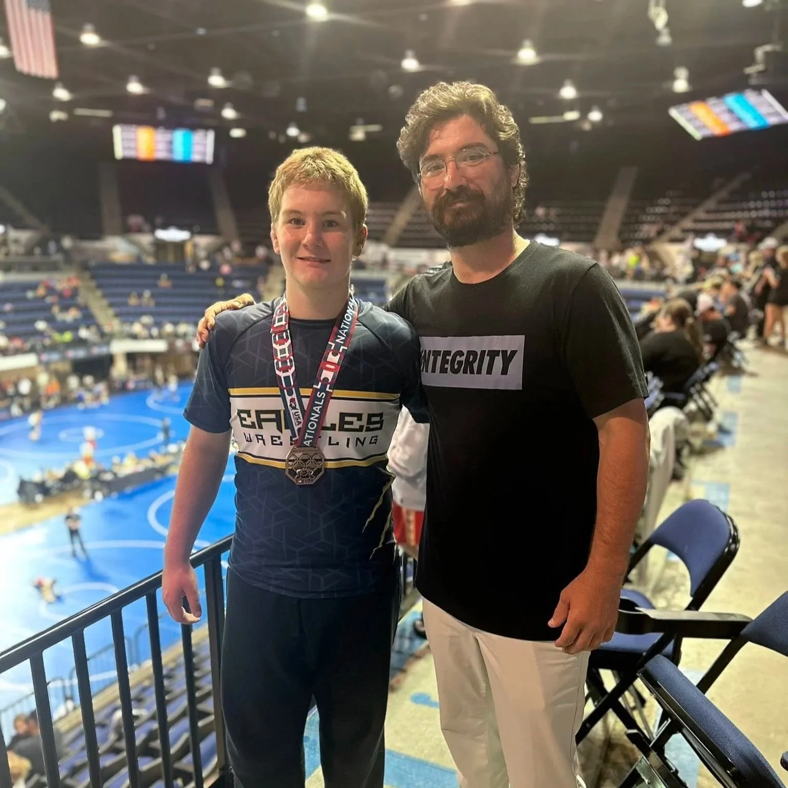 A young male athlete wearing a medal and a team shirt with the words 'Eagles Wrestling' poses with an older male in casual attire at a sports arena with a wrestling mat in the background.