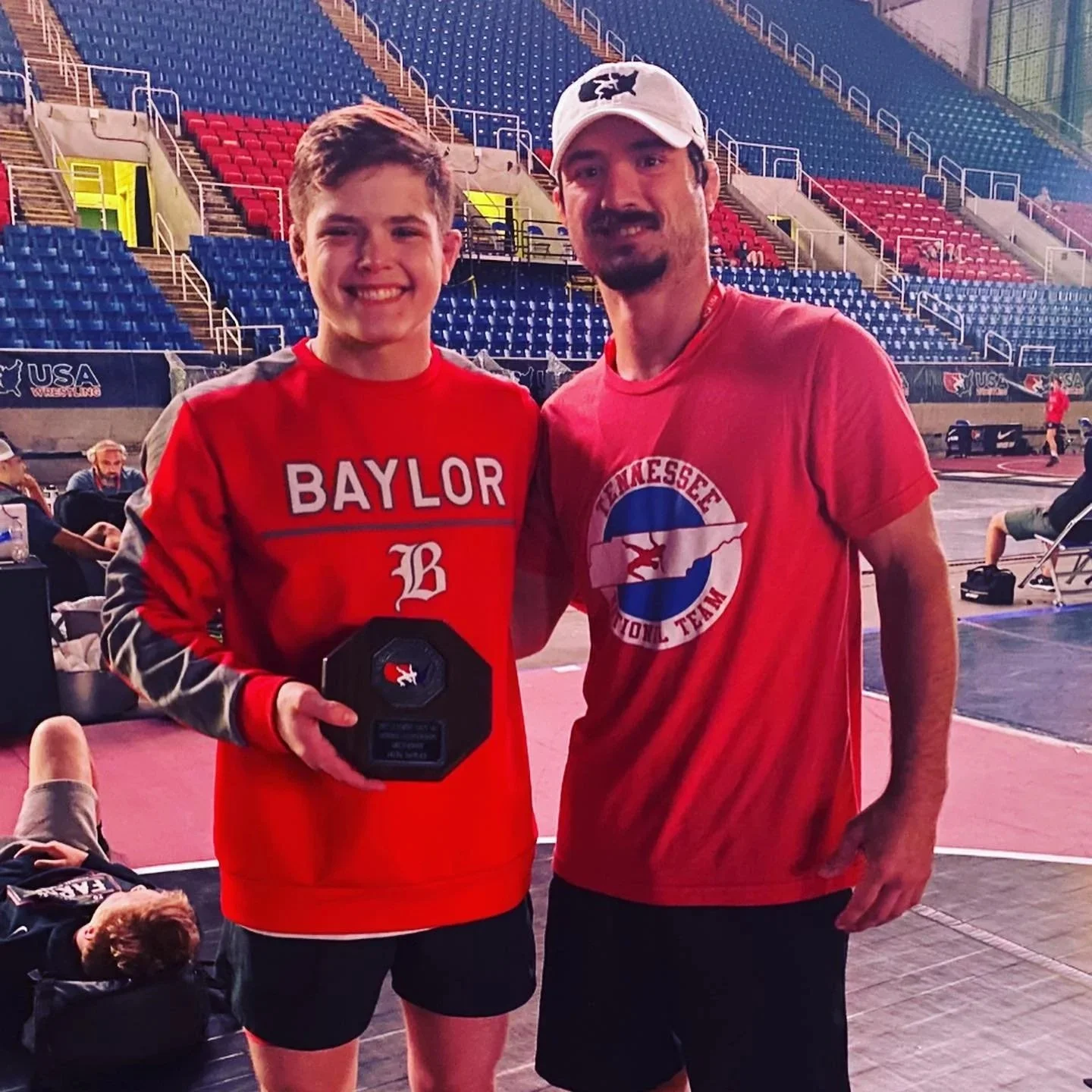 A young man in a red Baylor sweatshirt holding a trophy, standing next to an older man in a red Tennessee shirt inside a stadium or sports arena, with empty seats in the background.