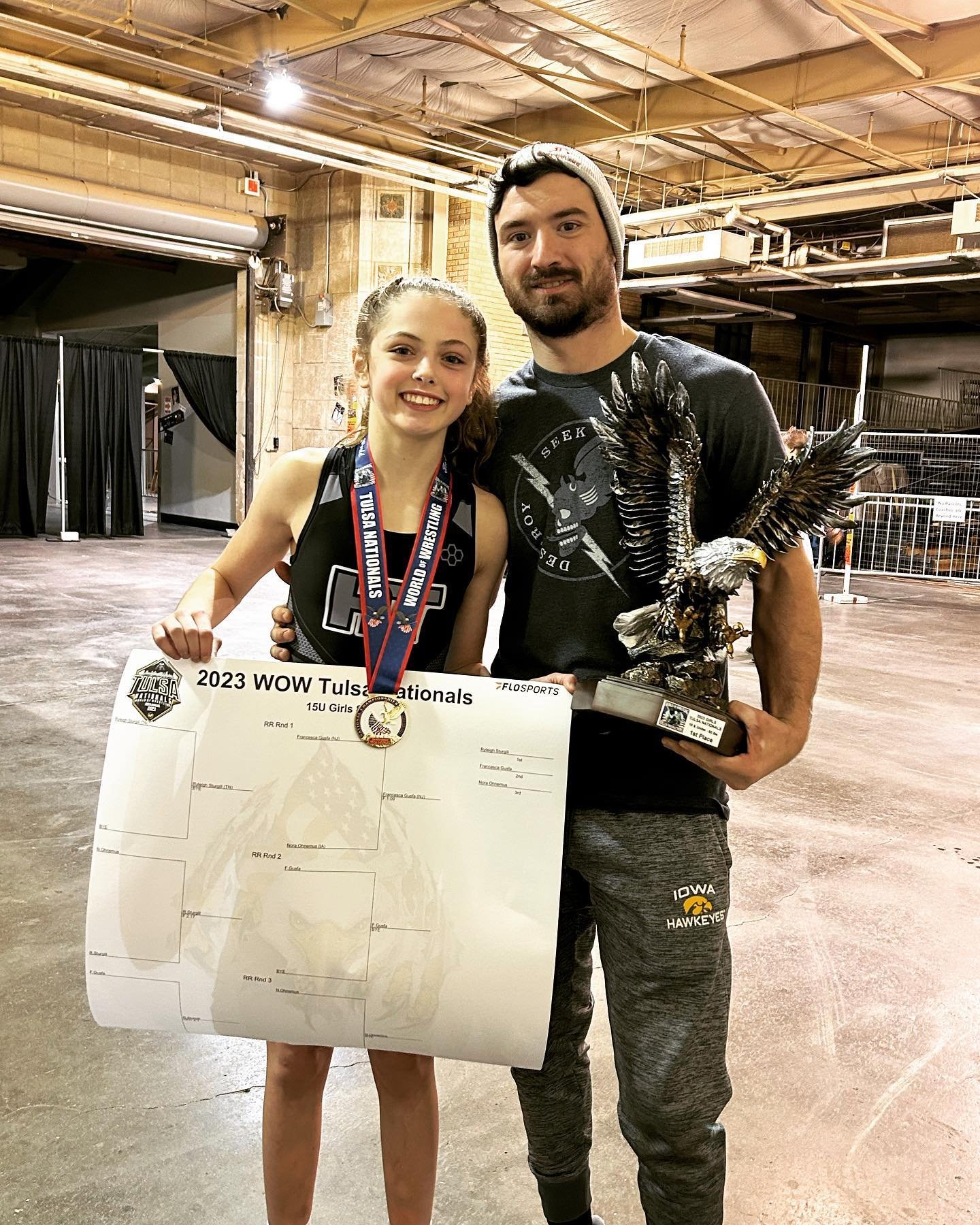 Young girl in a wrestling uniform holding a large tournament bracket sheet and wearing a medal, standing next to a man in casual sportswear holding a trophy of an eagle with outstretched wings, inside a gym or sports arena.