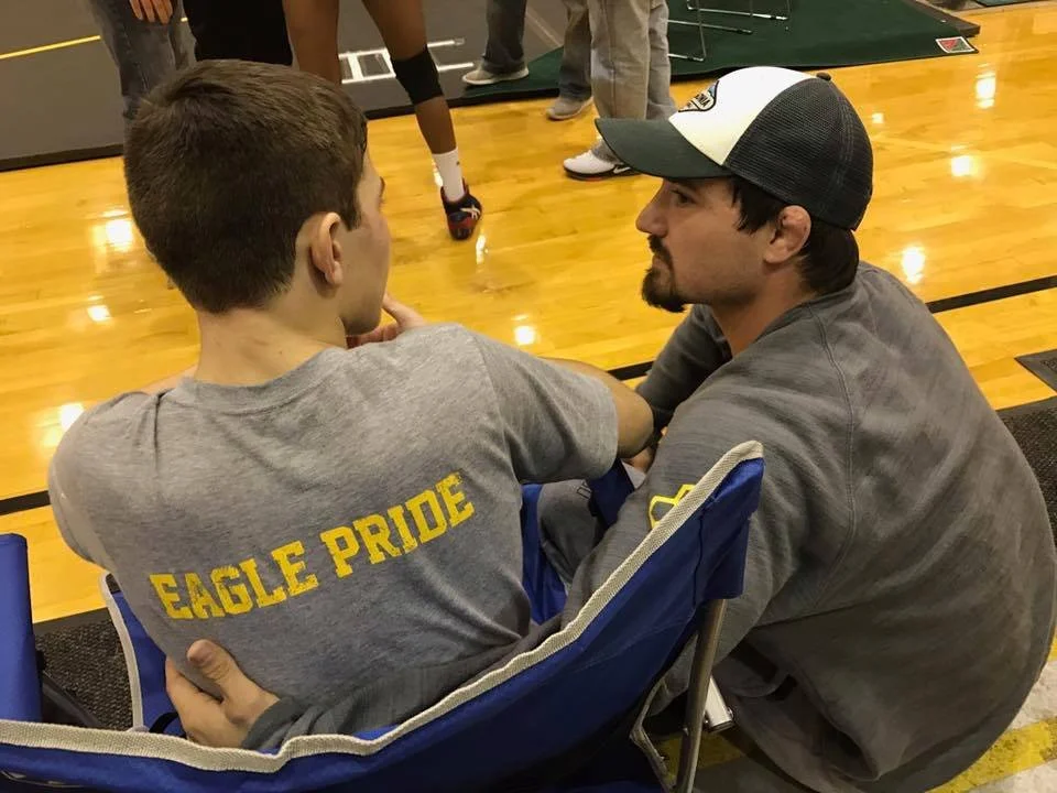 A young man wearing a gray shirt with 'EAGLE PRIDE' on the back, sitting in a folding chair, is talking to an adult man wearing a gray hoodie and a baseball cap, at a gymnasium.