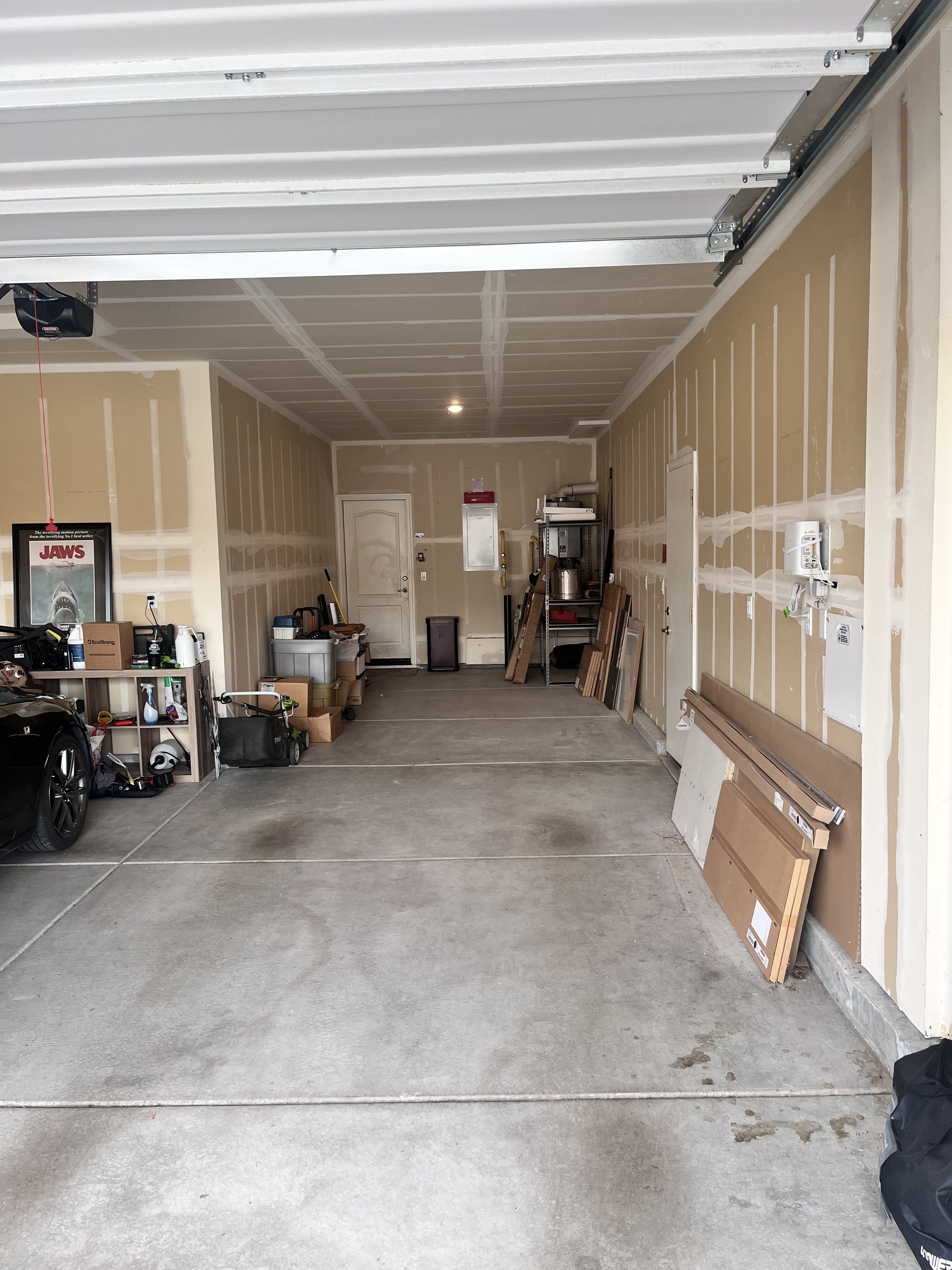 A clean garage with partially finished drywall, storage shelves, a car, and various boxes and tools. Garage door is open.