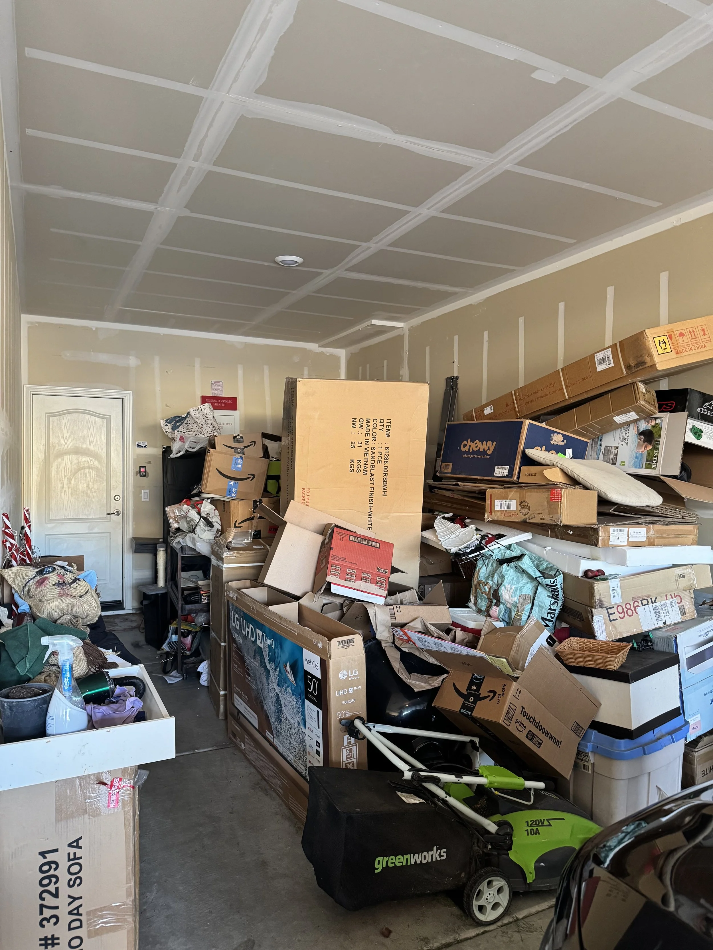 A cluttered garage filled with stacked boxes, bags, and household items, with a closed door on the left and a ceiling with visible drywall seams.