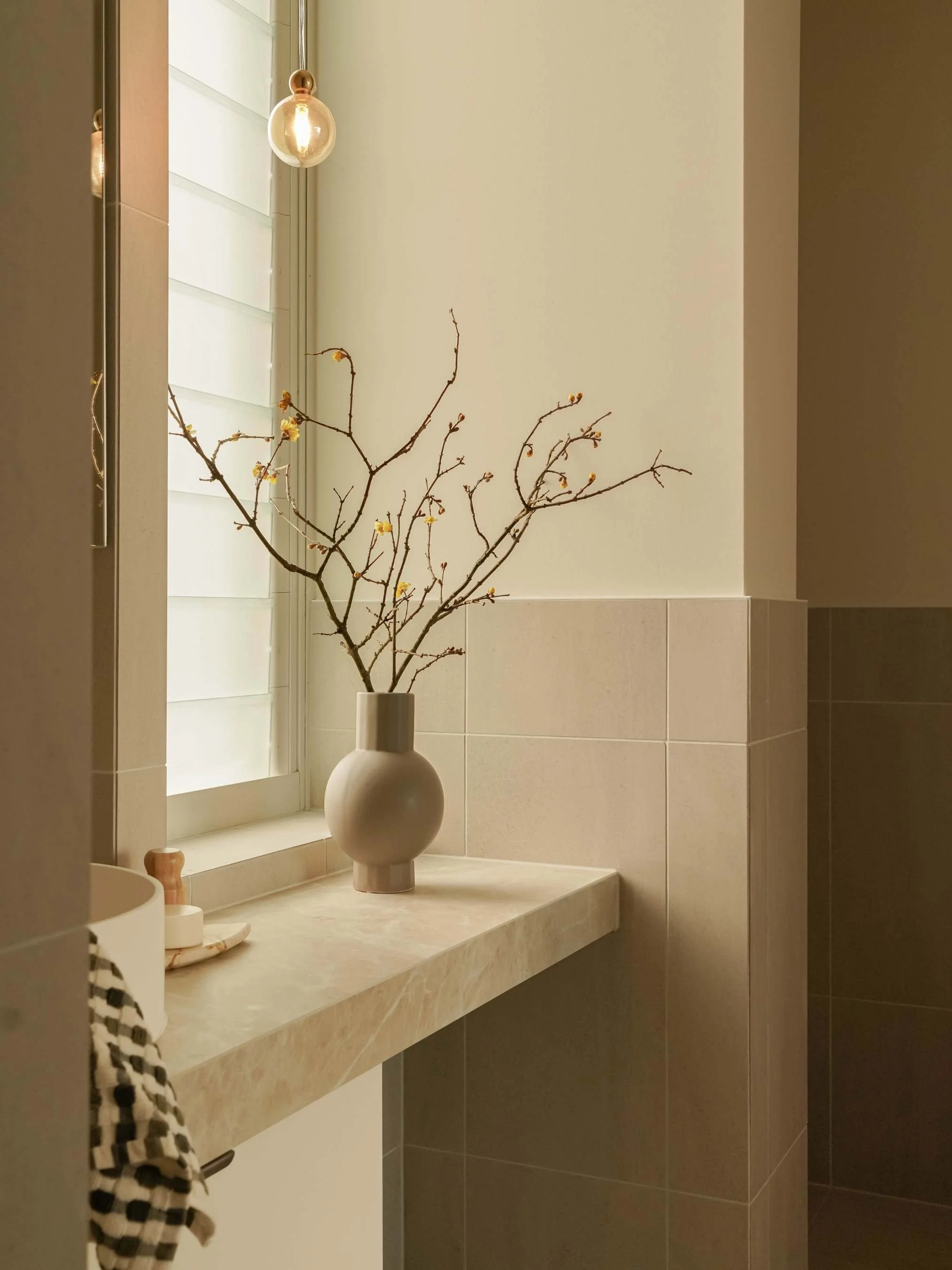 A beige marble countertop with a white vase containing bare branches with small yellow flowers, next to a window with white shutters, in a bathroom or similar interior space.
