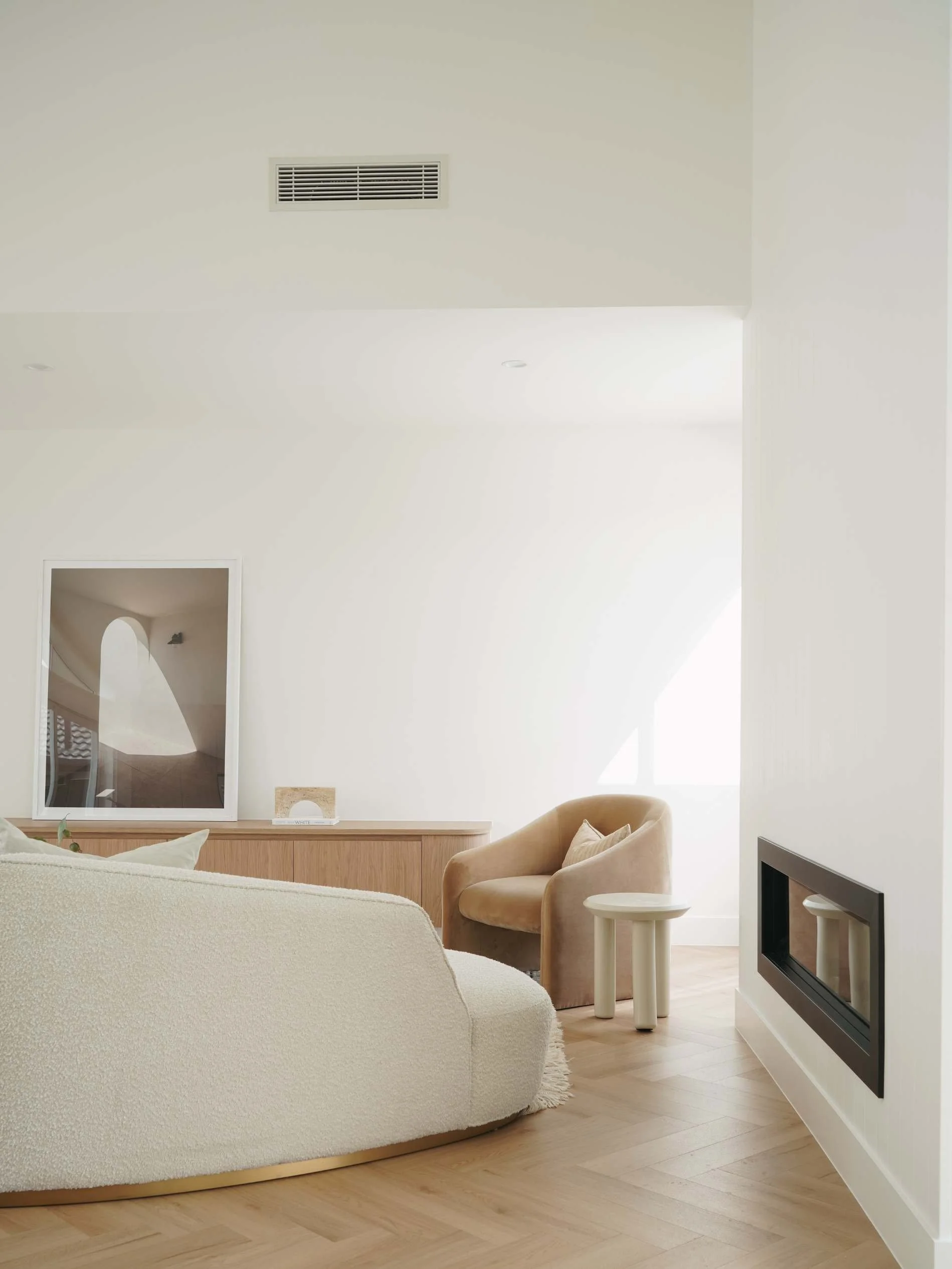 Minimalist Mediterranean living room with cream-colored curved sofa, tan armchair, small white side table, wood floor, and large window letting in natural light in Ostani House