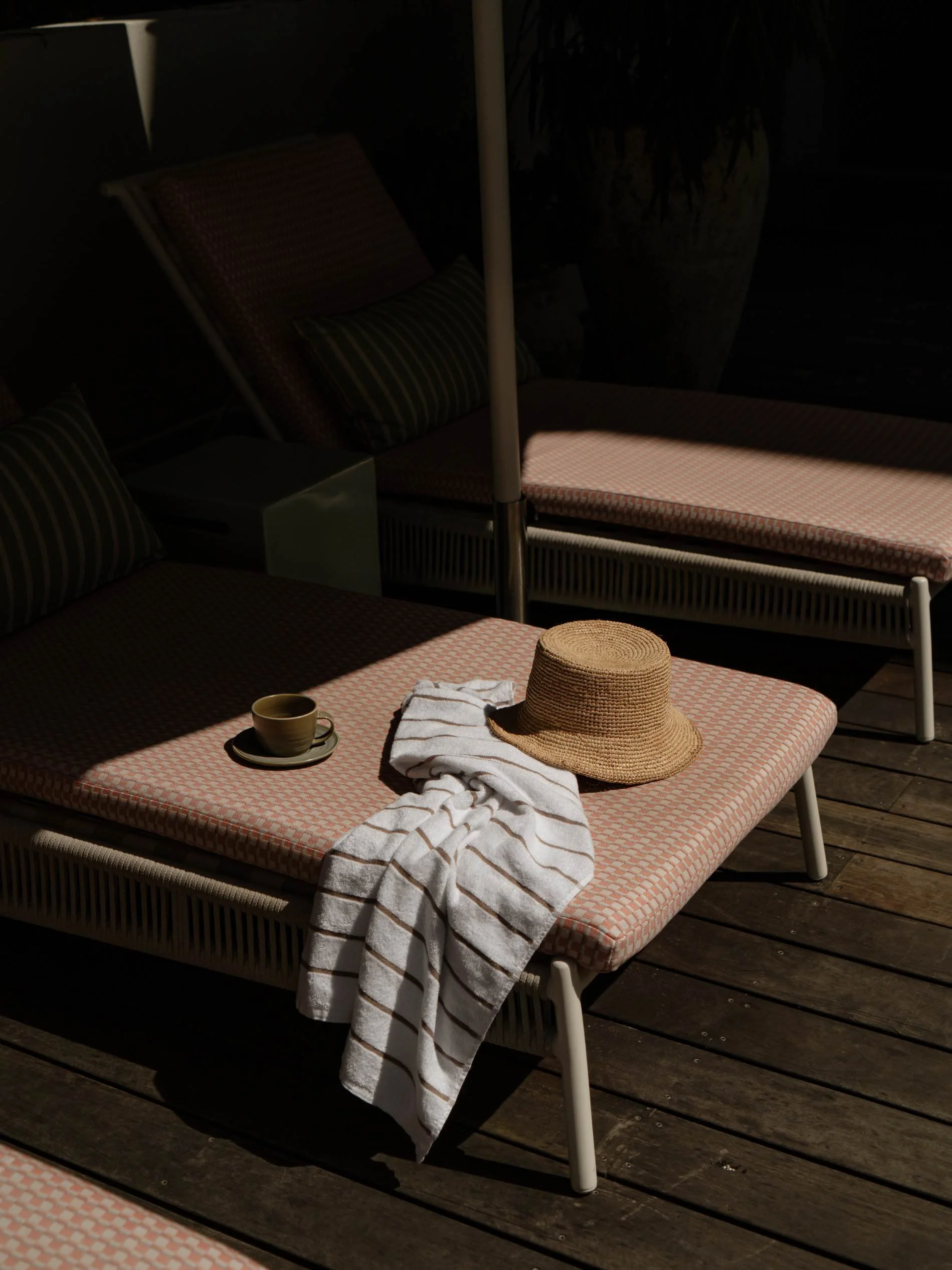 Beach lounge chairs with striped cushions, a straw hat, a towel, and a coffee cup on a wooden deck, partially shaded at Basq House Hotel in Byron Bay