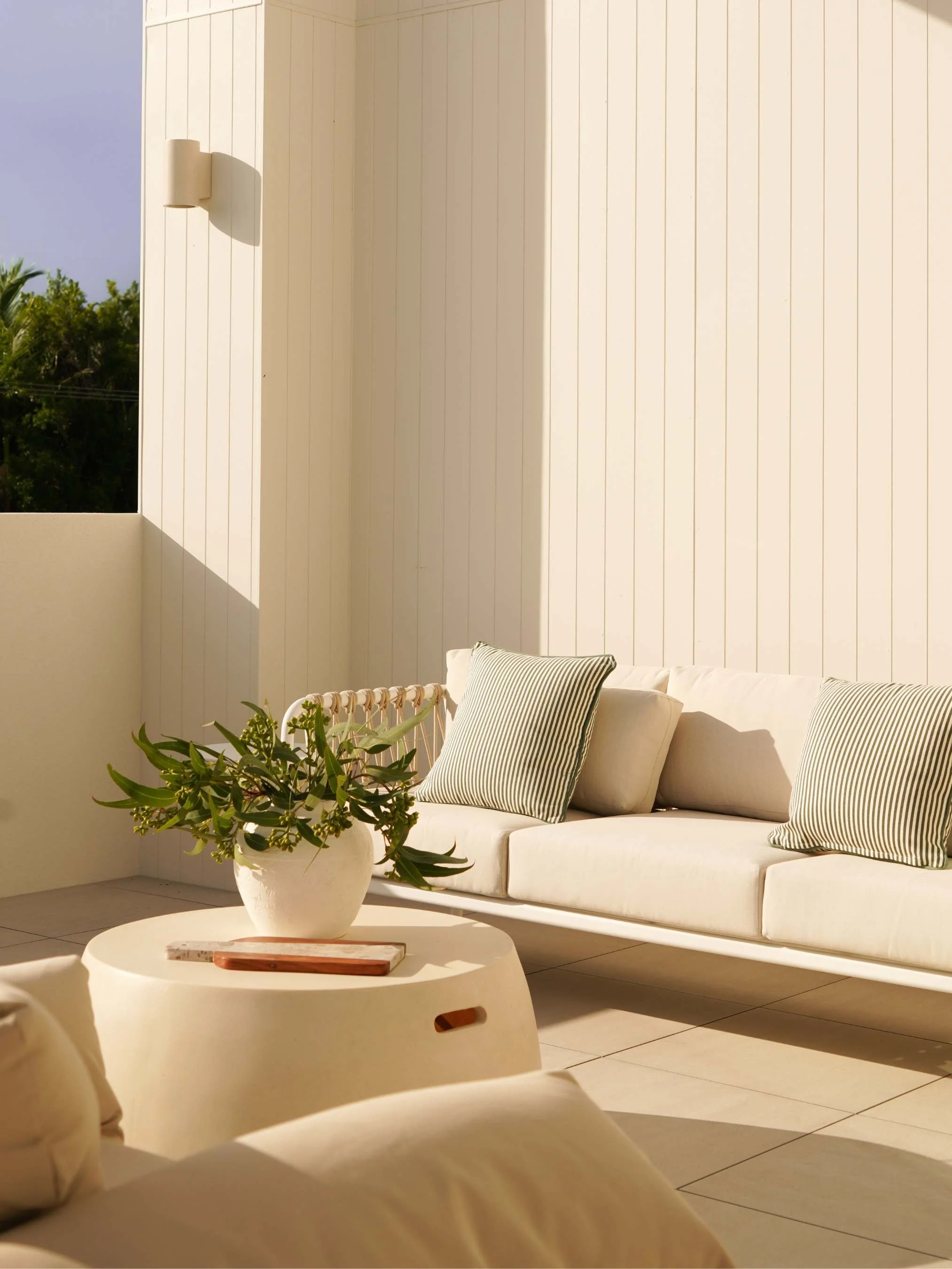 A modern patio area with a white outdoor sofa, striped pillows, a white round coffee table with green plant, and beige tiled flooring, with sunlight casting shadows on the wall in Noosa Heads, QLD