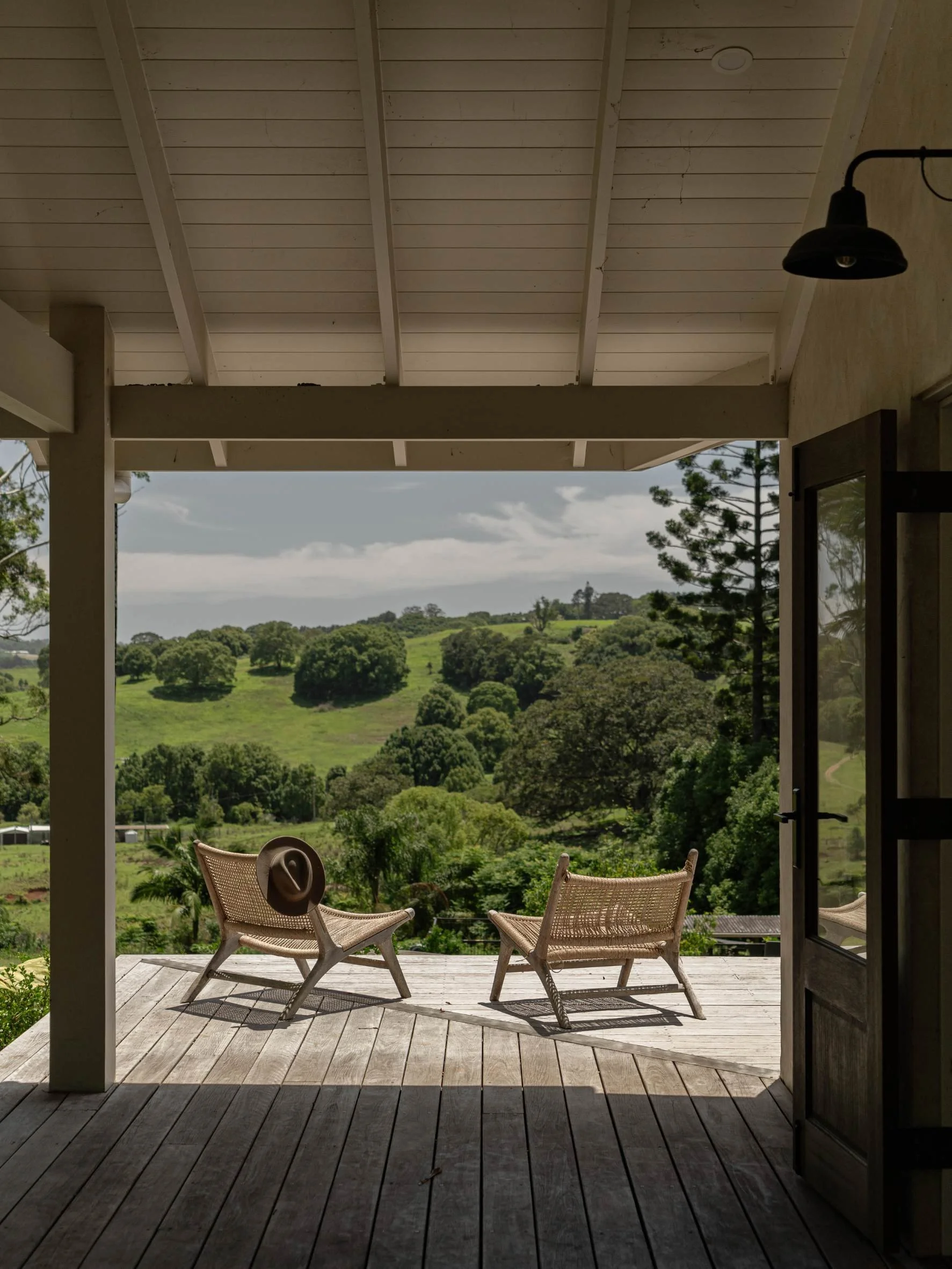 View from a shaded porch with two wicker chairs facing lush green hills and trees under a partly cloudy sky.