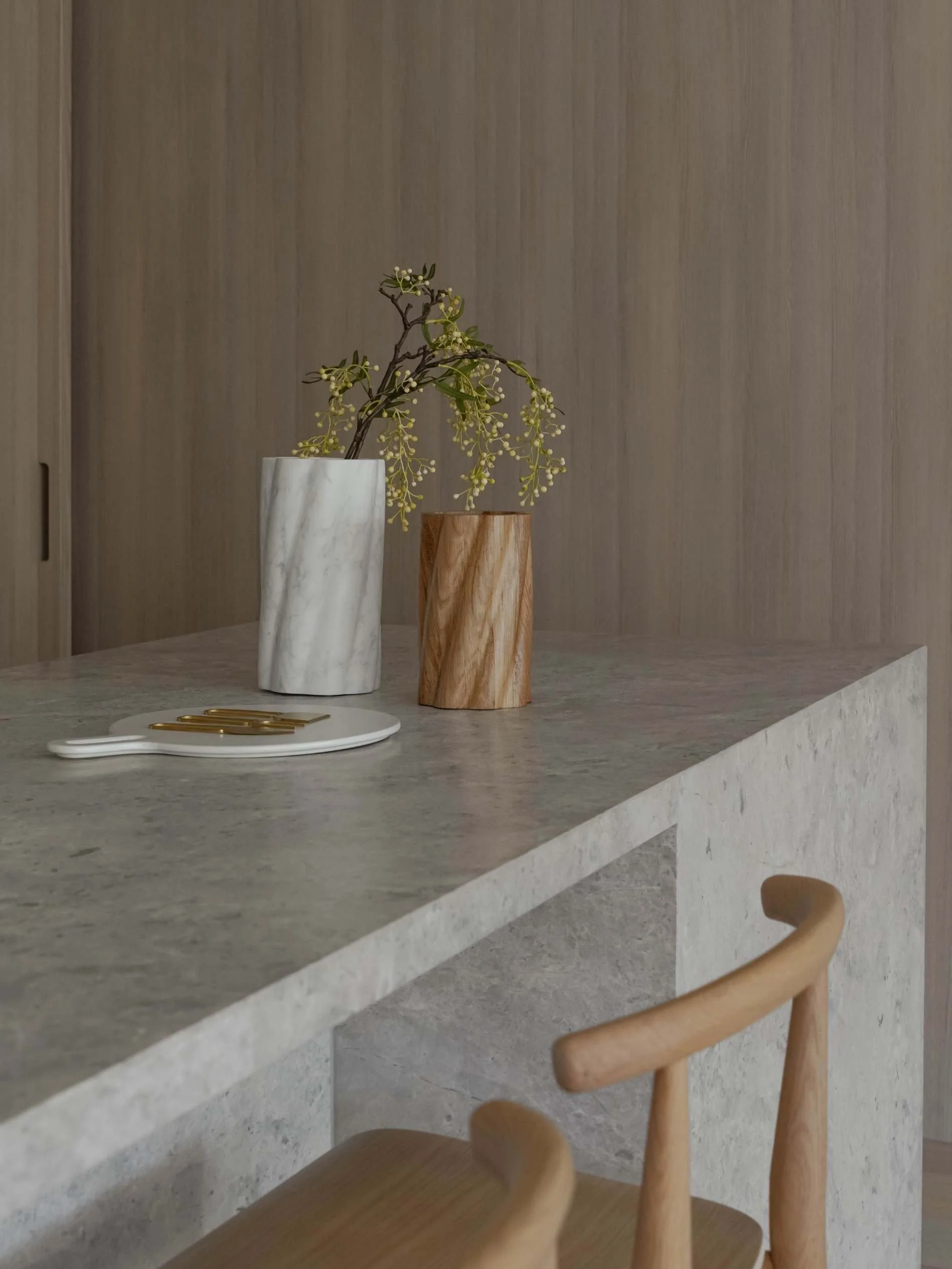 A kitchen countertop with two vases holding branches, one white marble and one wood, and a small white tray with gold cutlery.