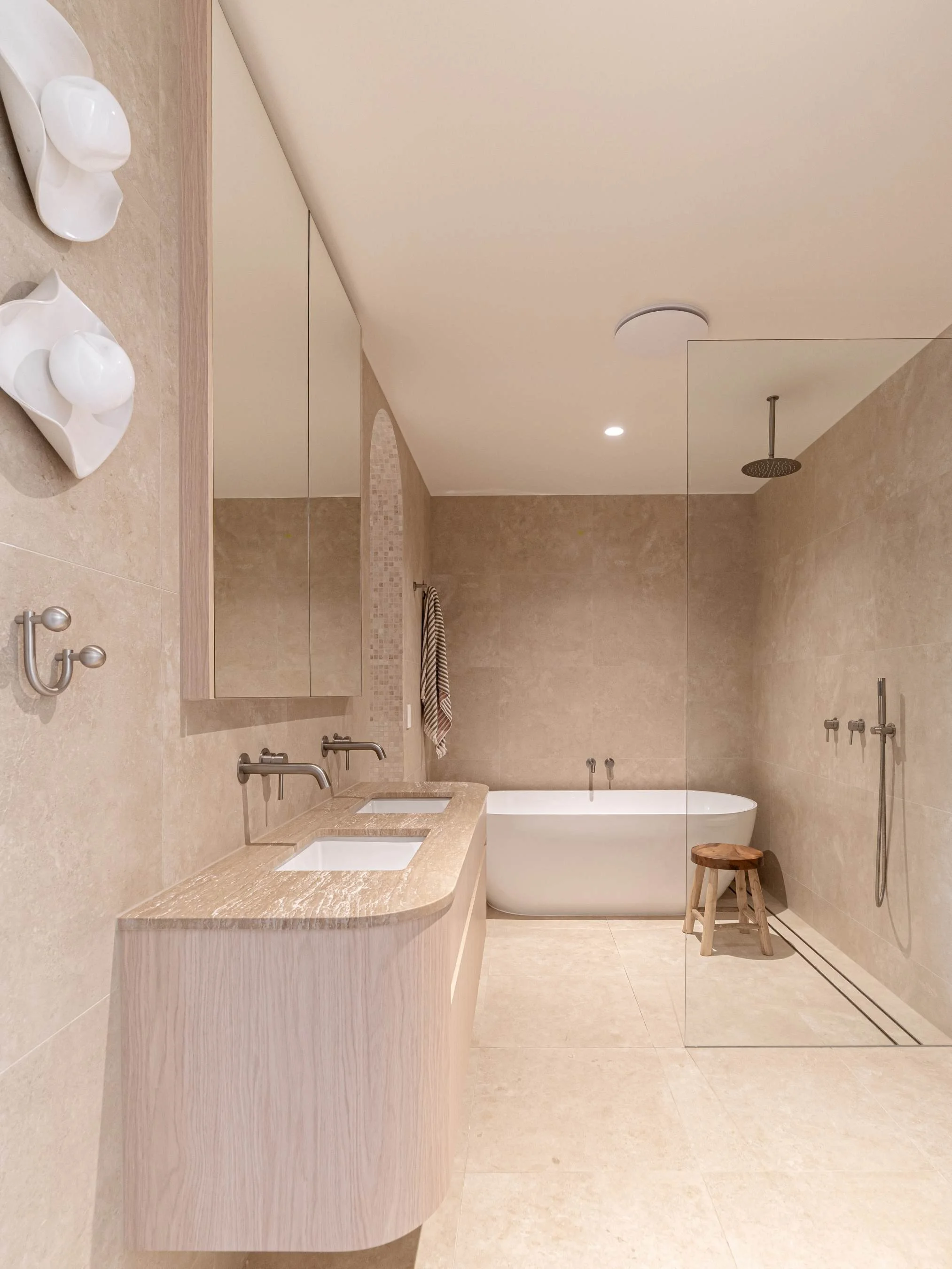 A modern bathroom with beige tiled walls and floors, a double sink vanity with marble countertop, a mirror, a bathtub, a small wooden stool, and a walk-in shower area with a hand-held showerhead and overhead rain shower.