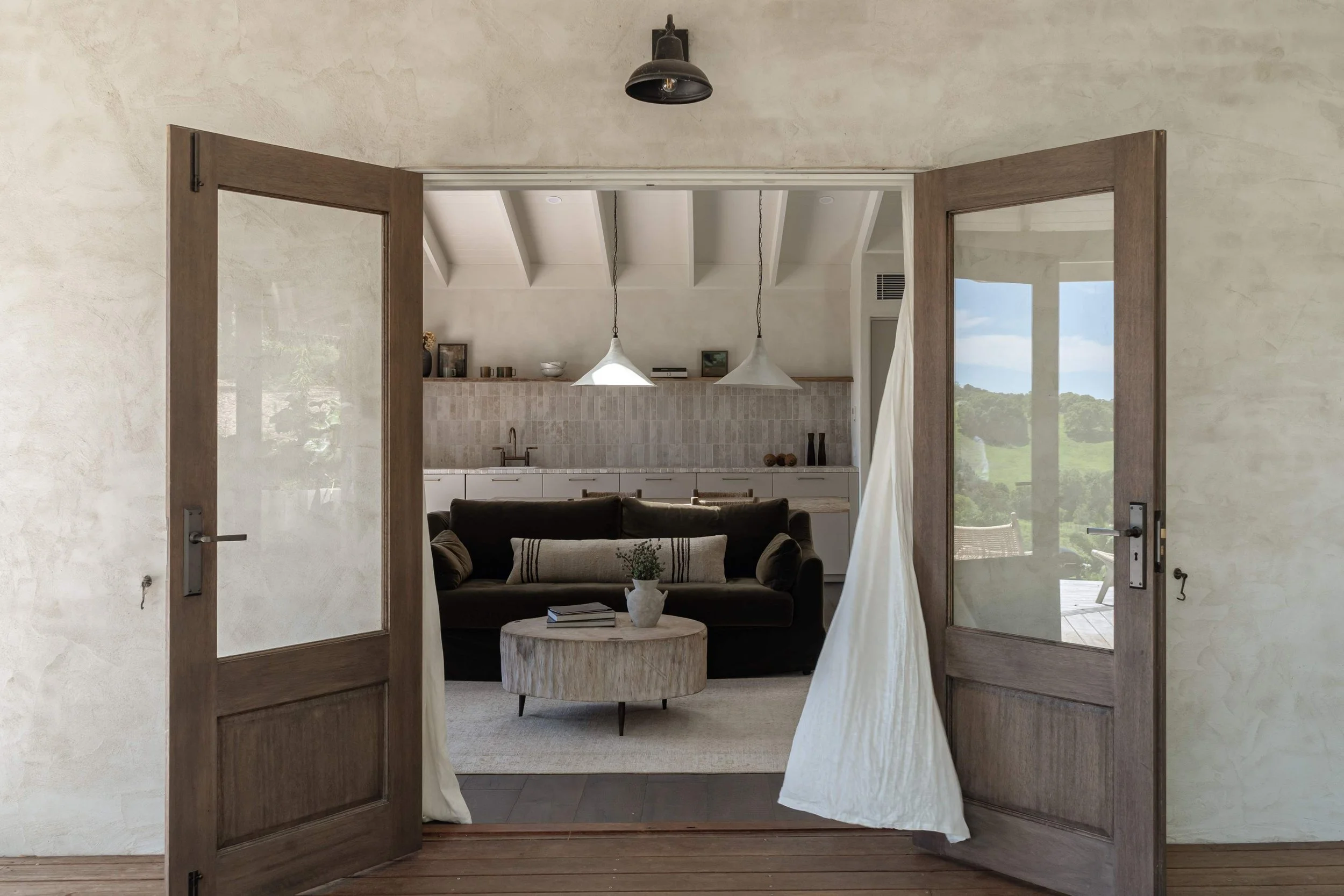 Open double French doors leading to a living room with a sofa, coffee table, and kitchen area with pendant lights, on a bright day outside. Featuring Hunter Cabin in Byron Bay