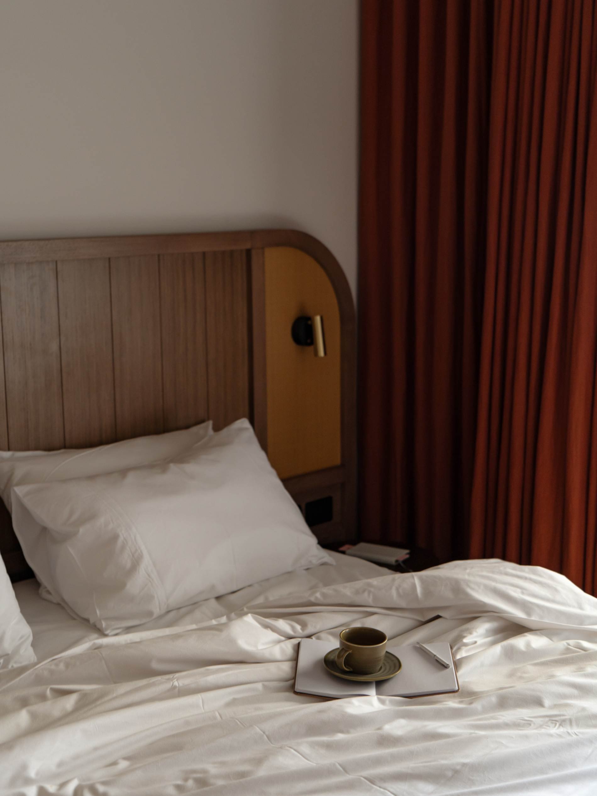 A neatly made hotel bed with white linens and pillows, a tray with a coffee cup and saucer, a notebook, and a pen on top. The headboard is wooden with a modern design and a reading light attached to it. A brown curtain is partially visible on the right side.
