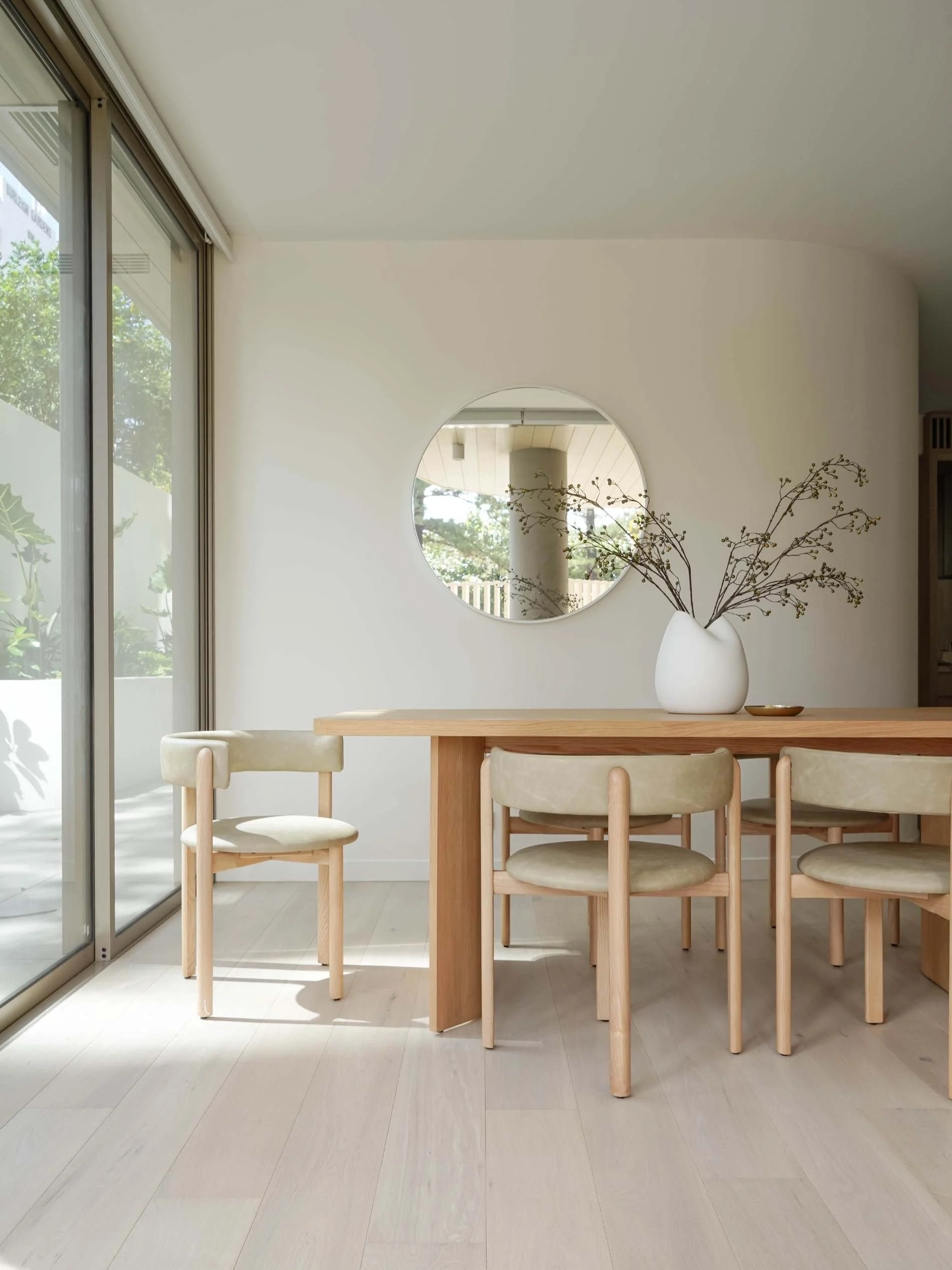 Minimalist dining area with wooden table, beige upholstered chairs, a white vase with branches, and a round mirror on the white wall at Florence, Mosaic Developers in Burleigh Heads