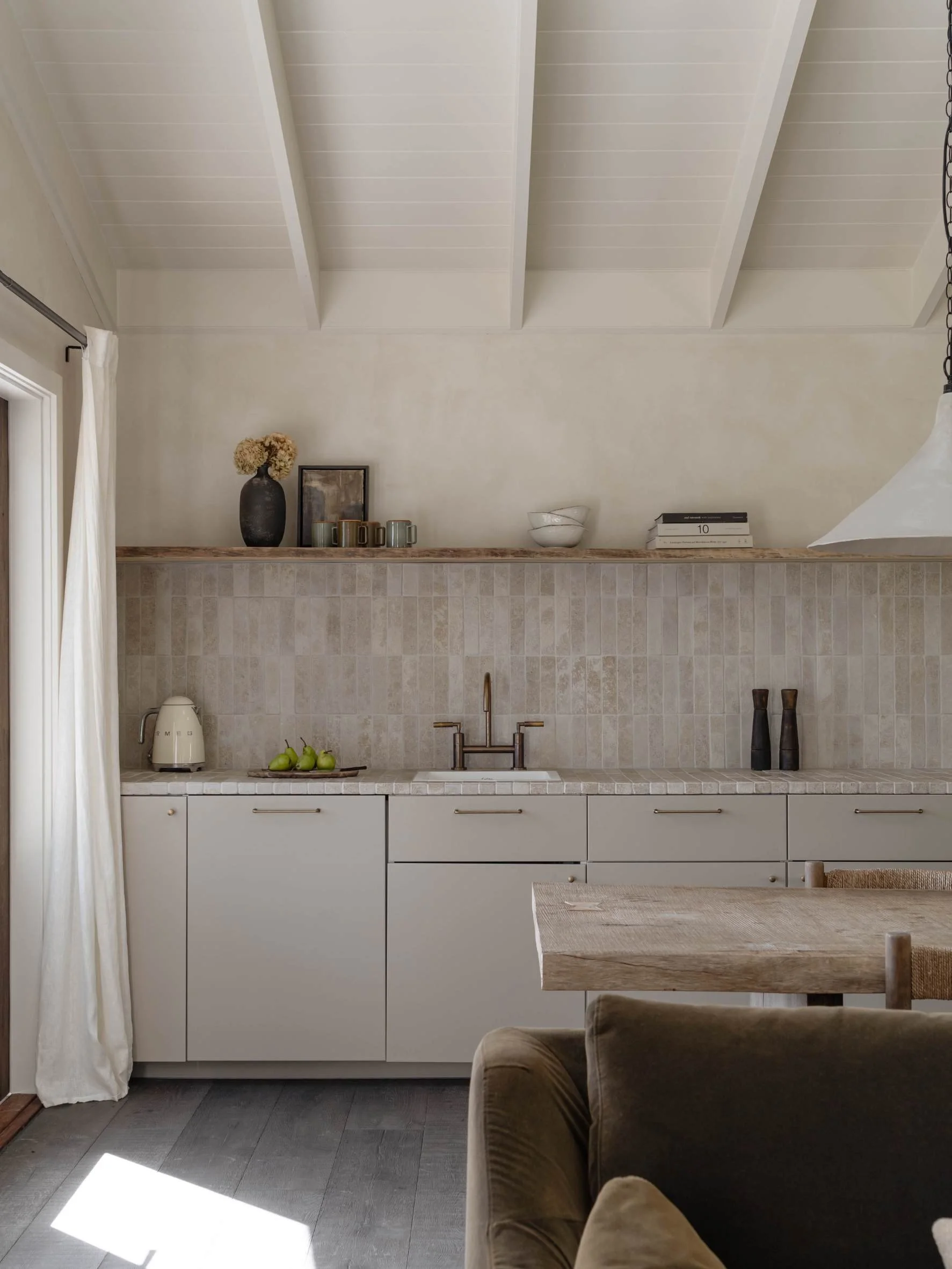 A minimalist kitchen with white cabinets, a wooden countertop, and beige tiled backsplash. Small appliances and decorative items are on the counter and shelf, including a kettle, pears, and vases. A wooden dining table with a chair is partially visib