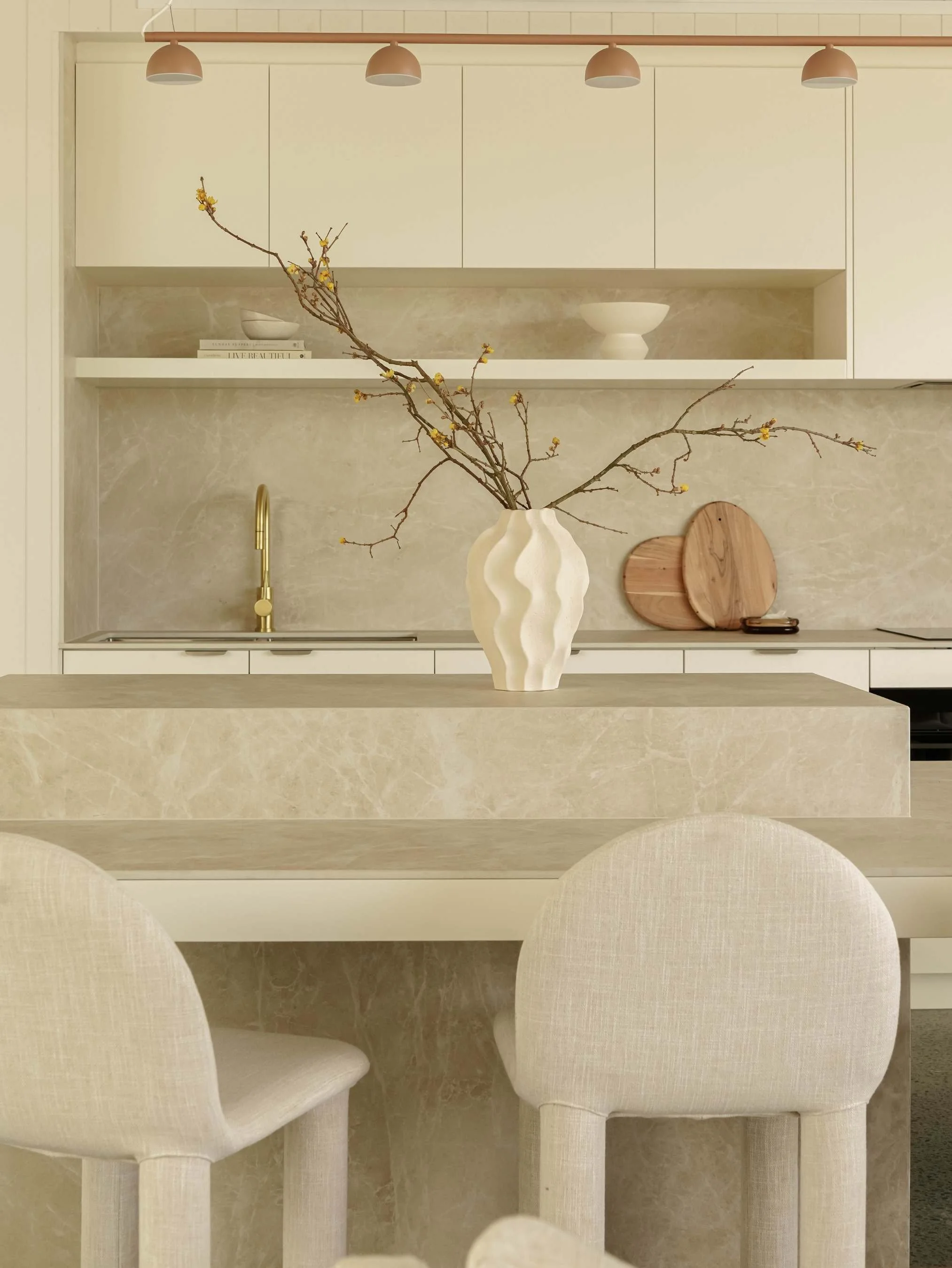 A minimalist kitchen with beige marble backsplash, beige countertops, and white cabinetry. A white vase with yellow flowering branches is on the kitchen island, with two round wooden cutting boards leaning against the wall. Beige chairs are in the fo