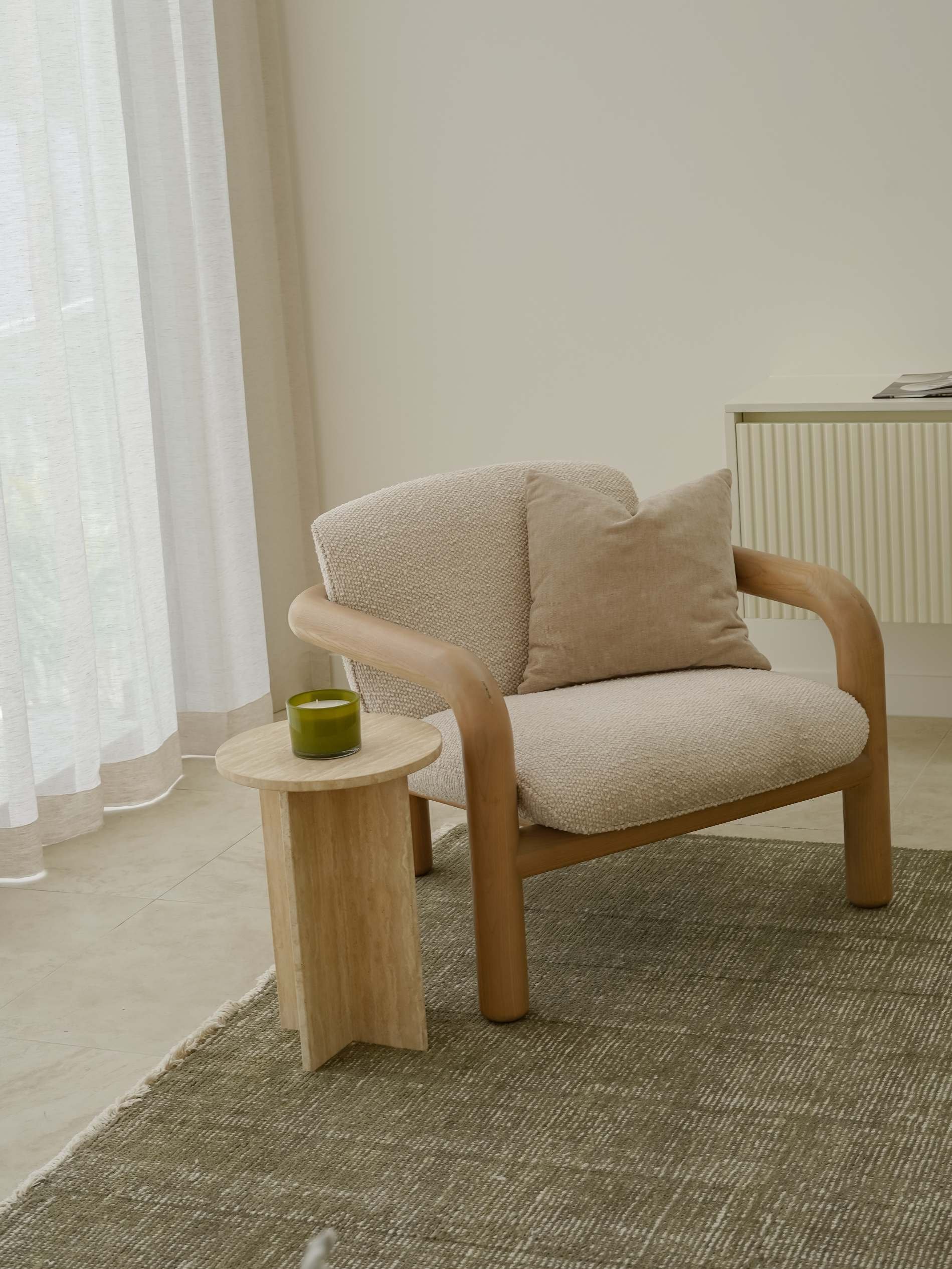 A modern living room corner with a beige armchair featuring wooden arms, a matching beige cushion, and a small round wooden side table with a green candle, next to a window with sheer white curtains and a white radiator.