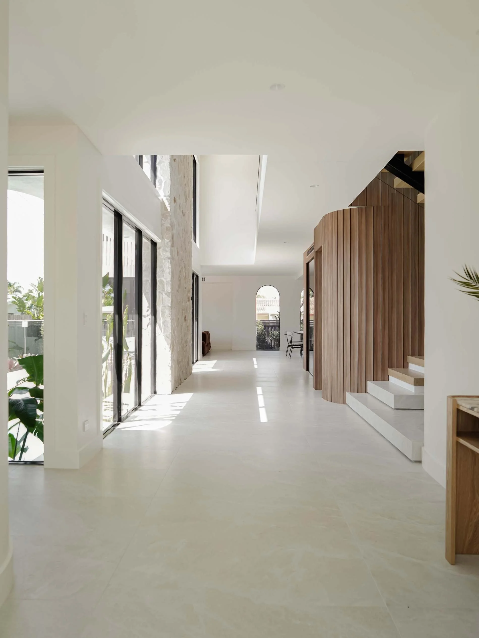 Bright modern interior with large sliding glass doors, white tiled floor, stone accent wall, wooden curved staircase, and a small dining area with chairs in the background in Savoy, Broadbeach Waters, QLD 
