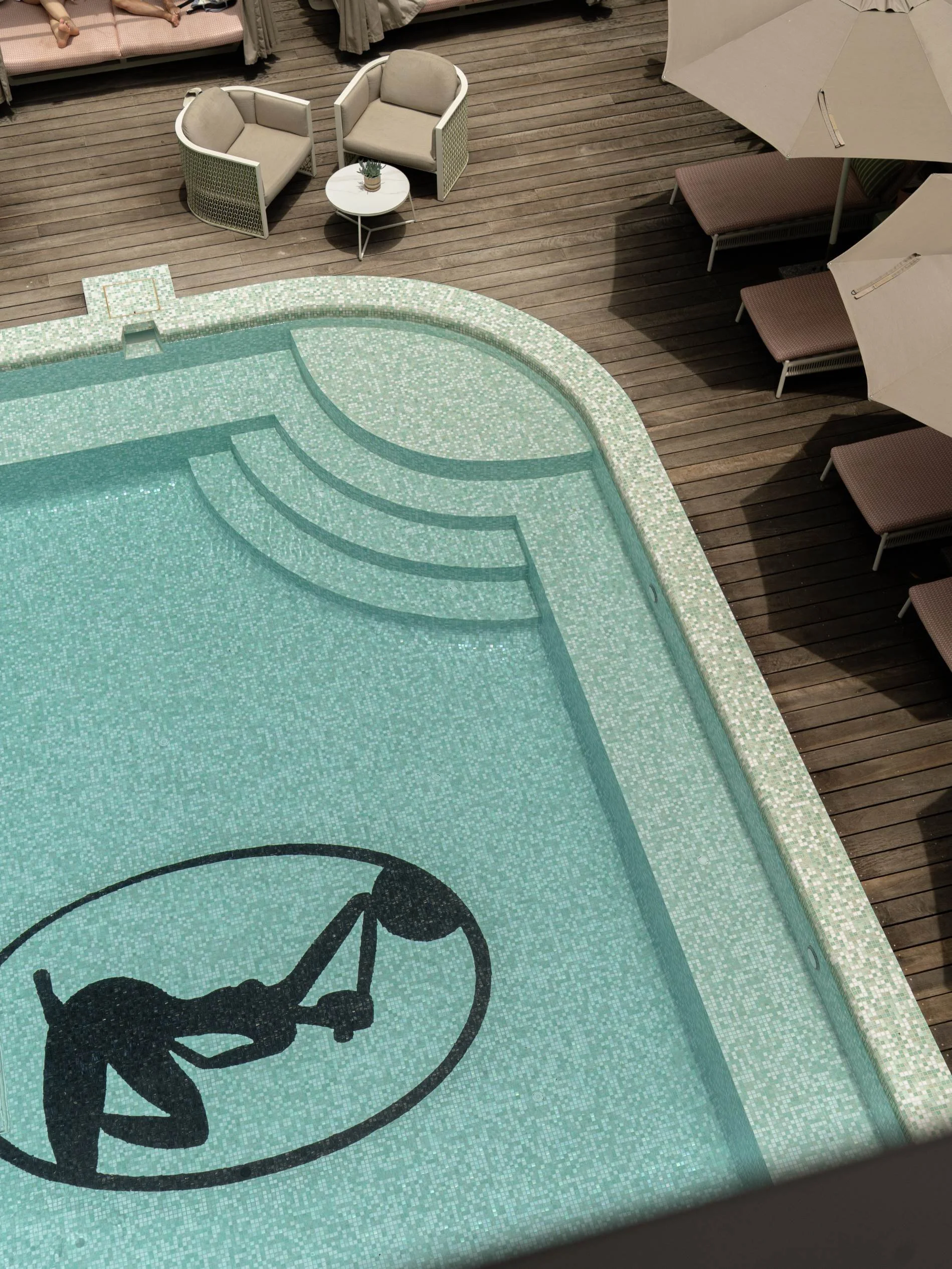 View of a swimming pool with a silhouette of a person holding a basketball and crouching inside a circle on the pool floor, surrounded by a wooden deck with lounge chairs, umbrellas, and seating area at Basq House Hotel in Byron Bay