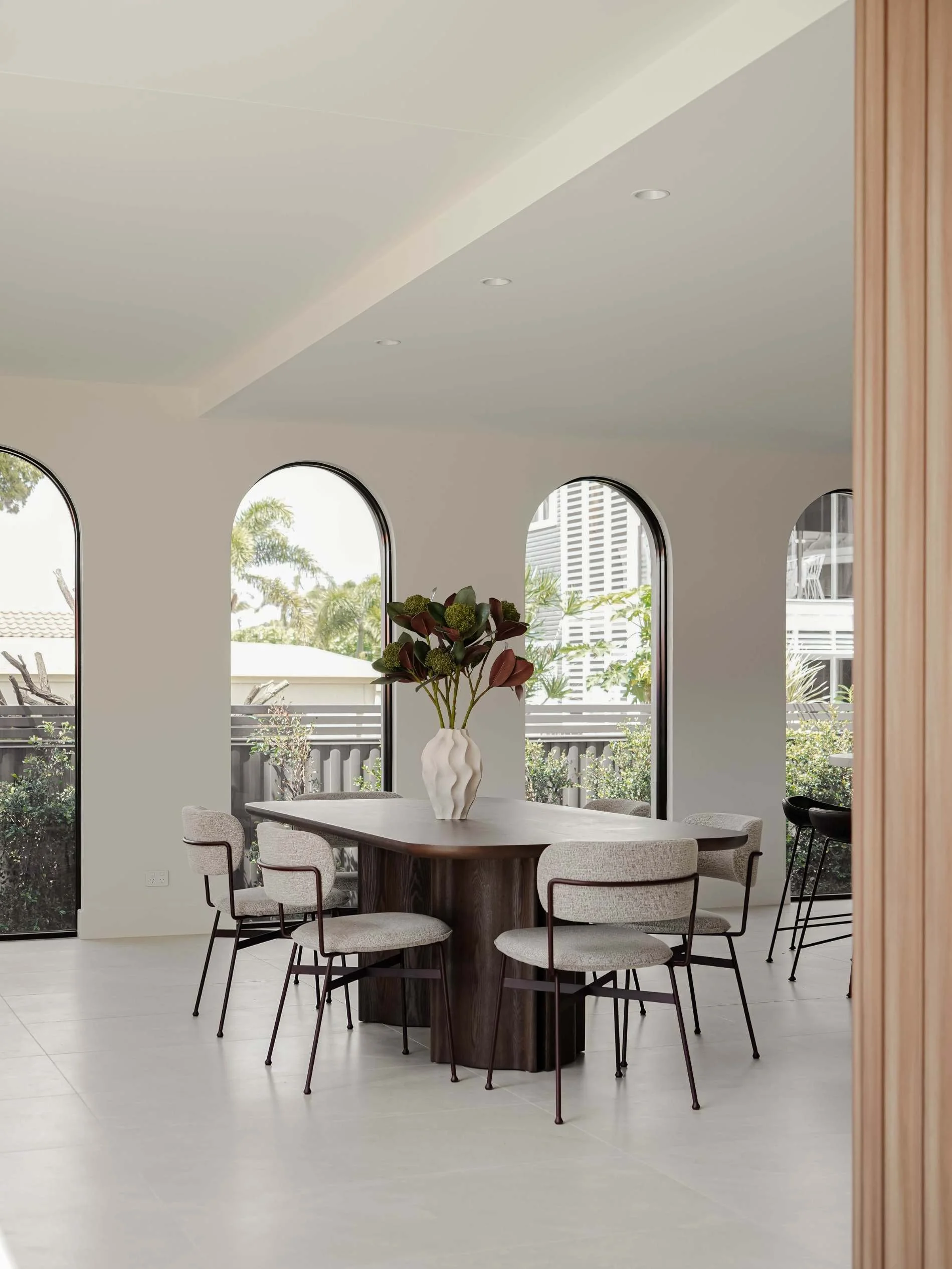 Modern dining room with a dark wooden oval table, surrounded by beige upholstered chairs with dark metal legs, a white textured vase with green and red plants, large arched windows, and neutral color palette in Savoy, Broadbeach Waters, QLD 