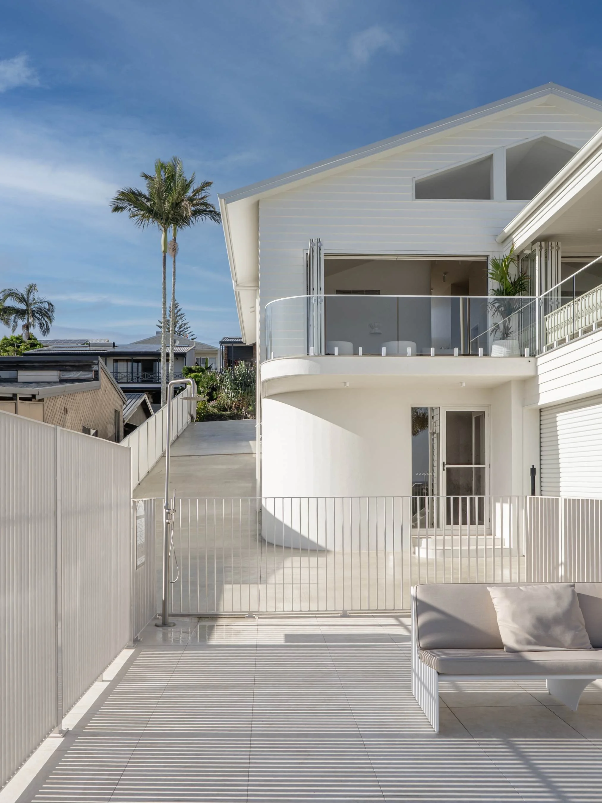Modern white two-story house with balcony, sliding glass doors, and outdoor patio featuring white benches and a fence, under a blue sky with palm trees in the background in the Ocean Ave Project in Tweed Heads, QLD