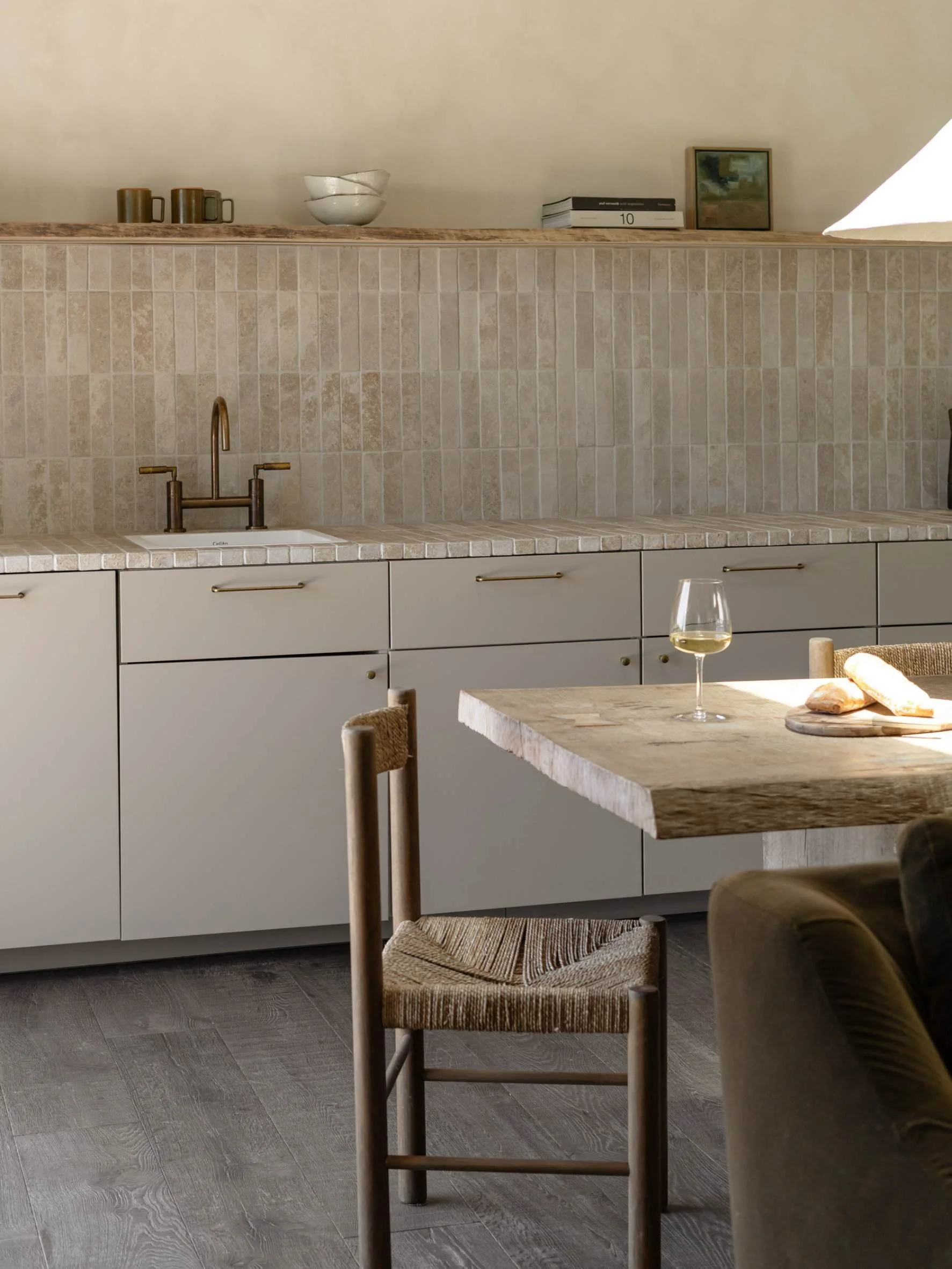 Modern kitchen with beige tile backsplash, gray cabinets, wooden dining table with a glass of white wine, bread, and a chair.