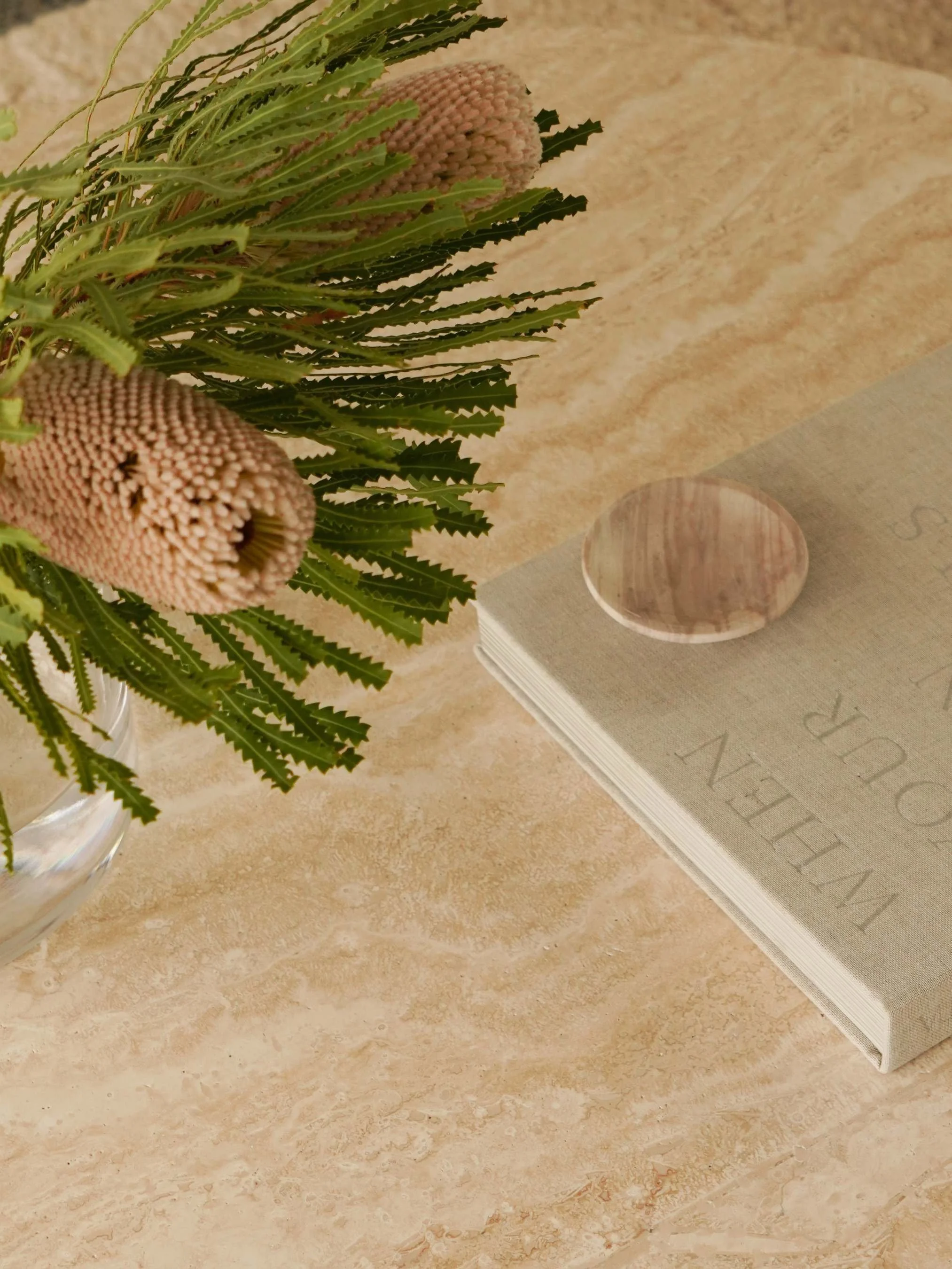 A beige marble table with a green book titled 'VOGUE MAY 2019' resting on it. There is a small round stone coaster on top of the book. A glass vase with pink flowers and green foliage is partially visible to the left of the frame.