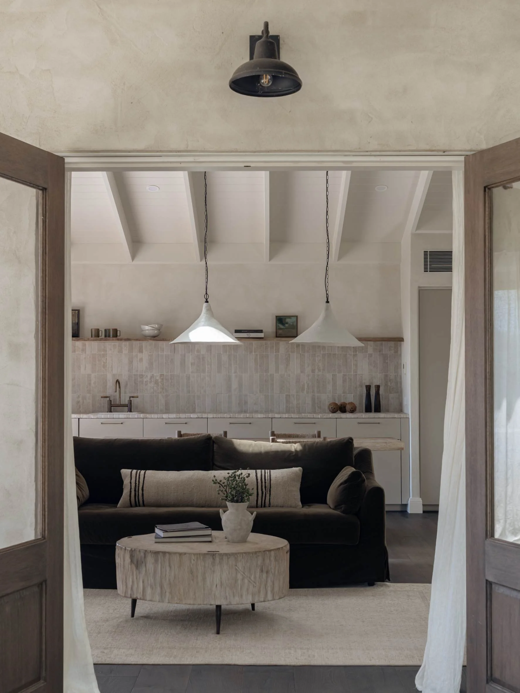 View into a living room with a black sofa, a wooden round coffee table with a potted plant and books, beige wall with white curtains, and a kitchen with white cabinets, beige tiled backsplash, and hanging pendant lights.