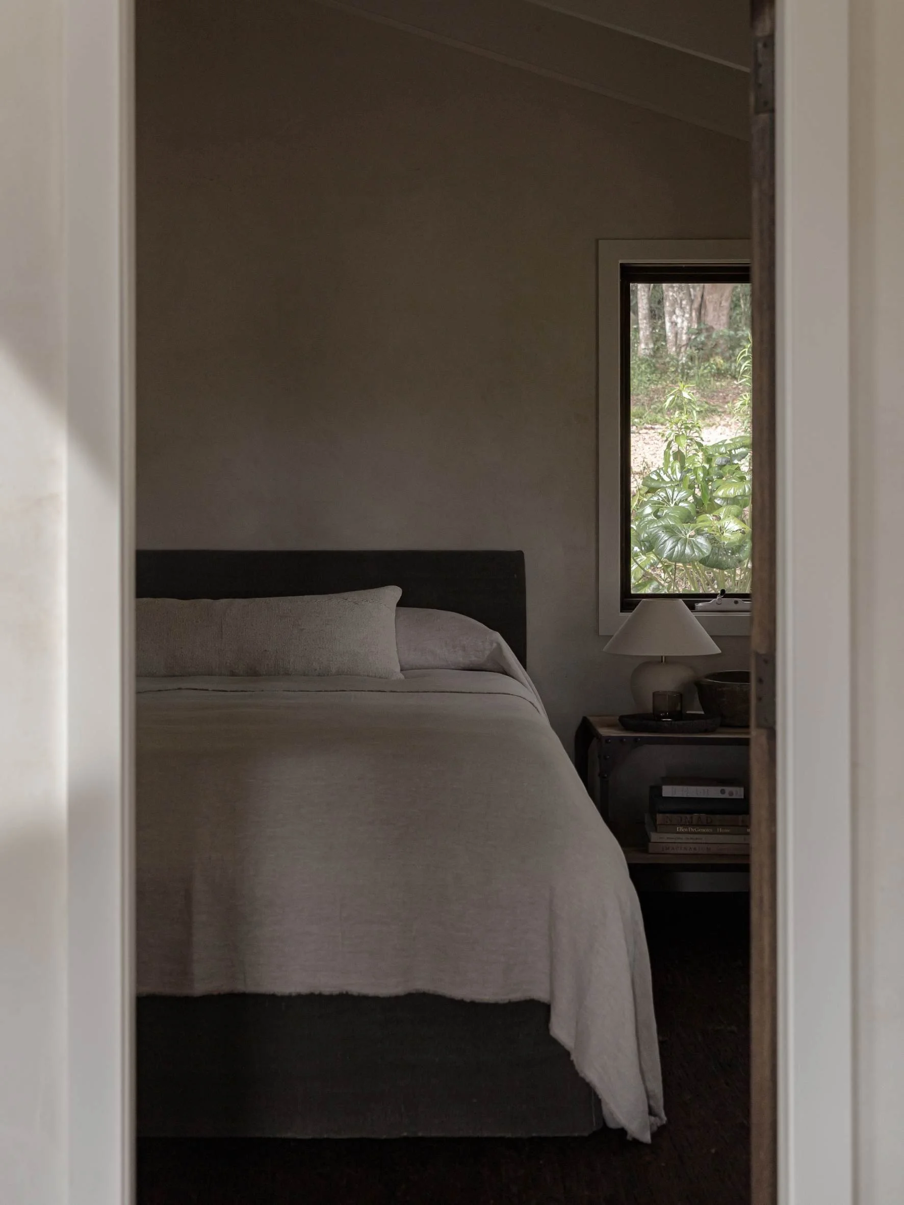 A cozy bedroom seen through a doorway, with a neatly made bed, a nightstand with a lamp, books, and decor, and a window showing greenery outside.