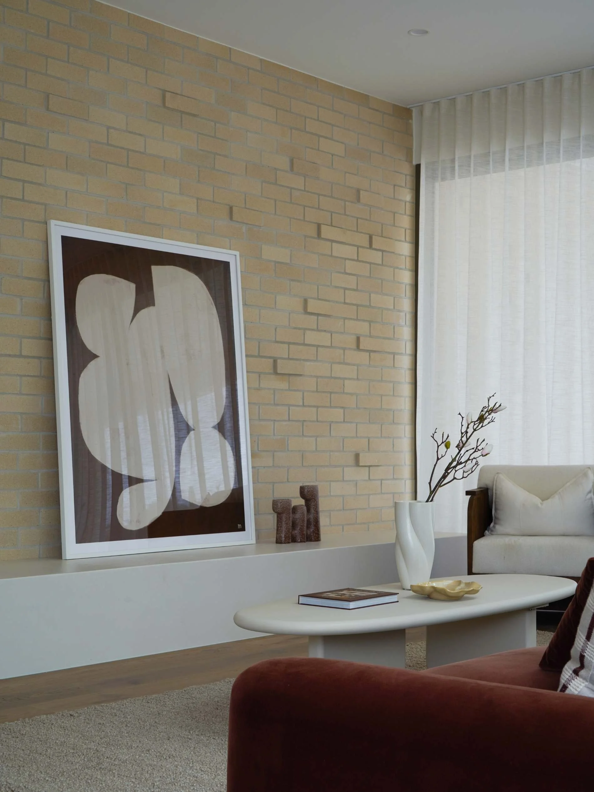 Mid century palm springs Living room with beige brick wall, a large framed abstract art piece leaning against wall, a white coffee table with a vase, a book, cream curtains, a beige sofa, and a cushioned chair in honeyeater drive burleigh waters