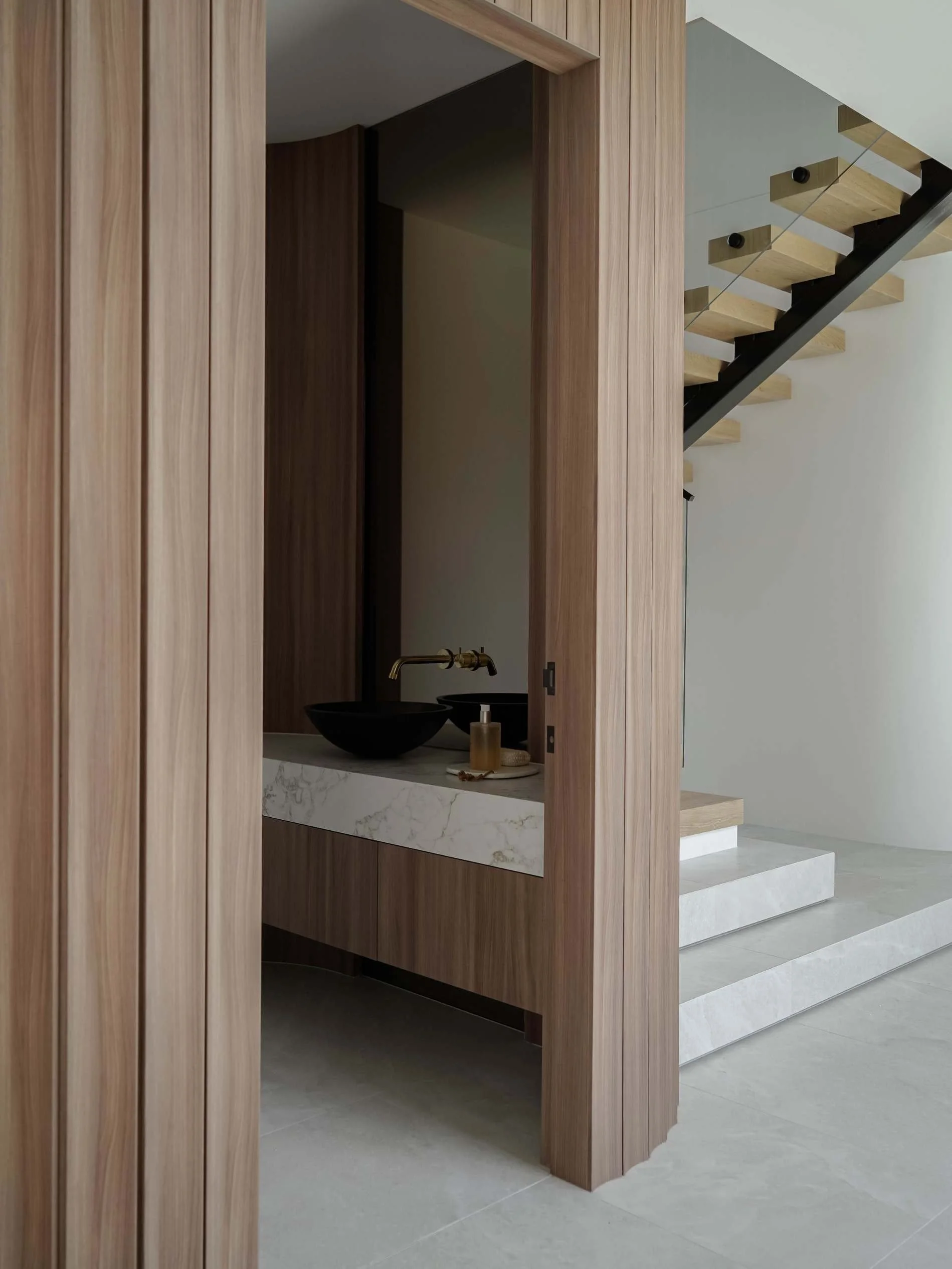 View of a modern interior featuring a staircase with wooden steps and a black railing, a small sink area with a black basin, a marble countertop, and a wooden wall.