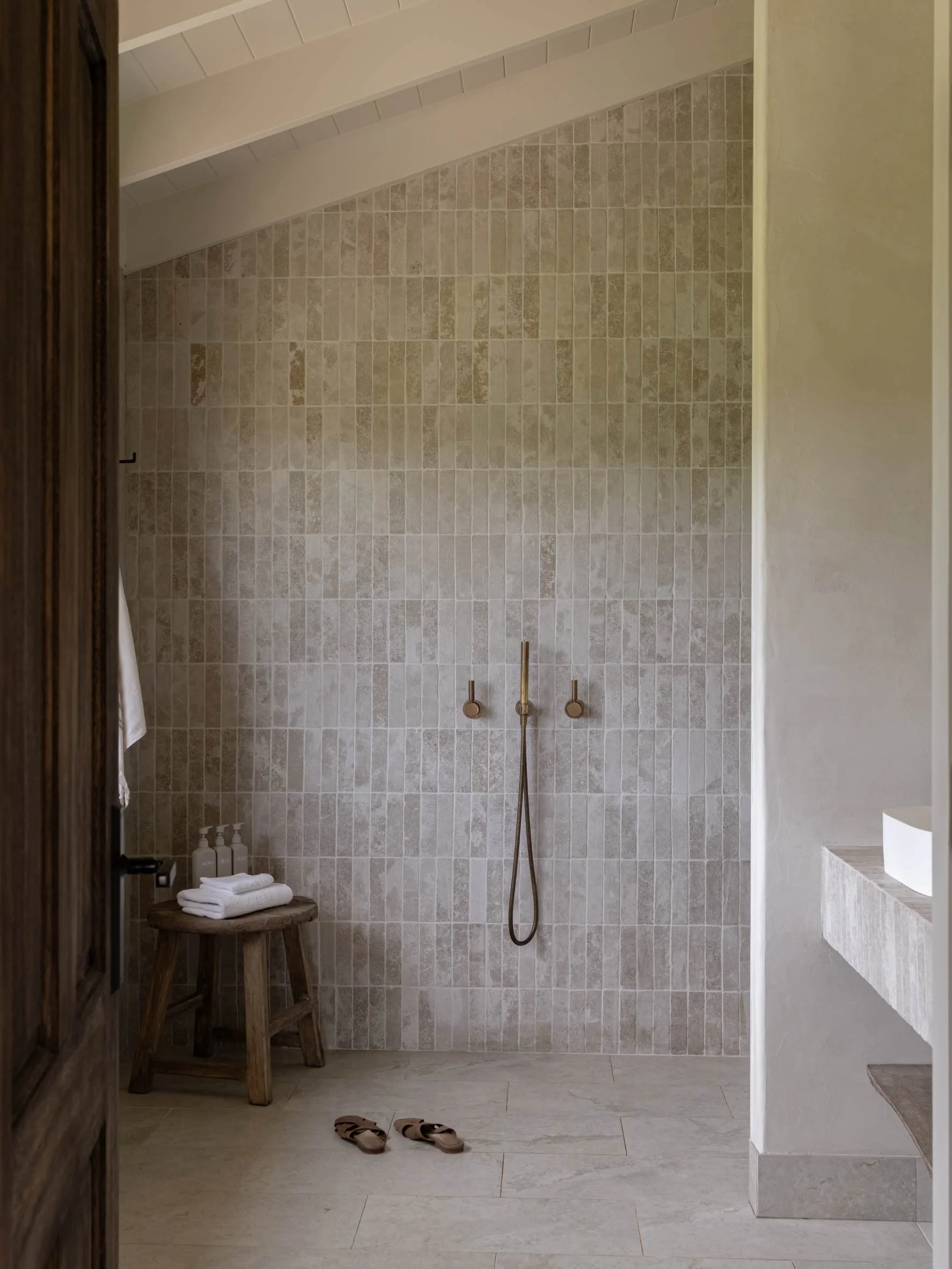 Minimalist bathroom with beige tiles, a wall-mounted showerhead, small wooden stool with towels, and a pair of slippers on the floor.