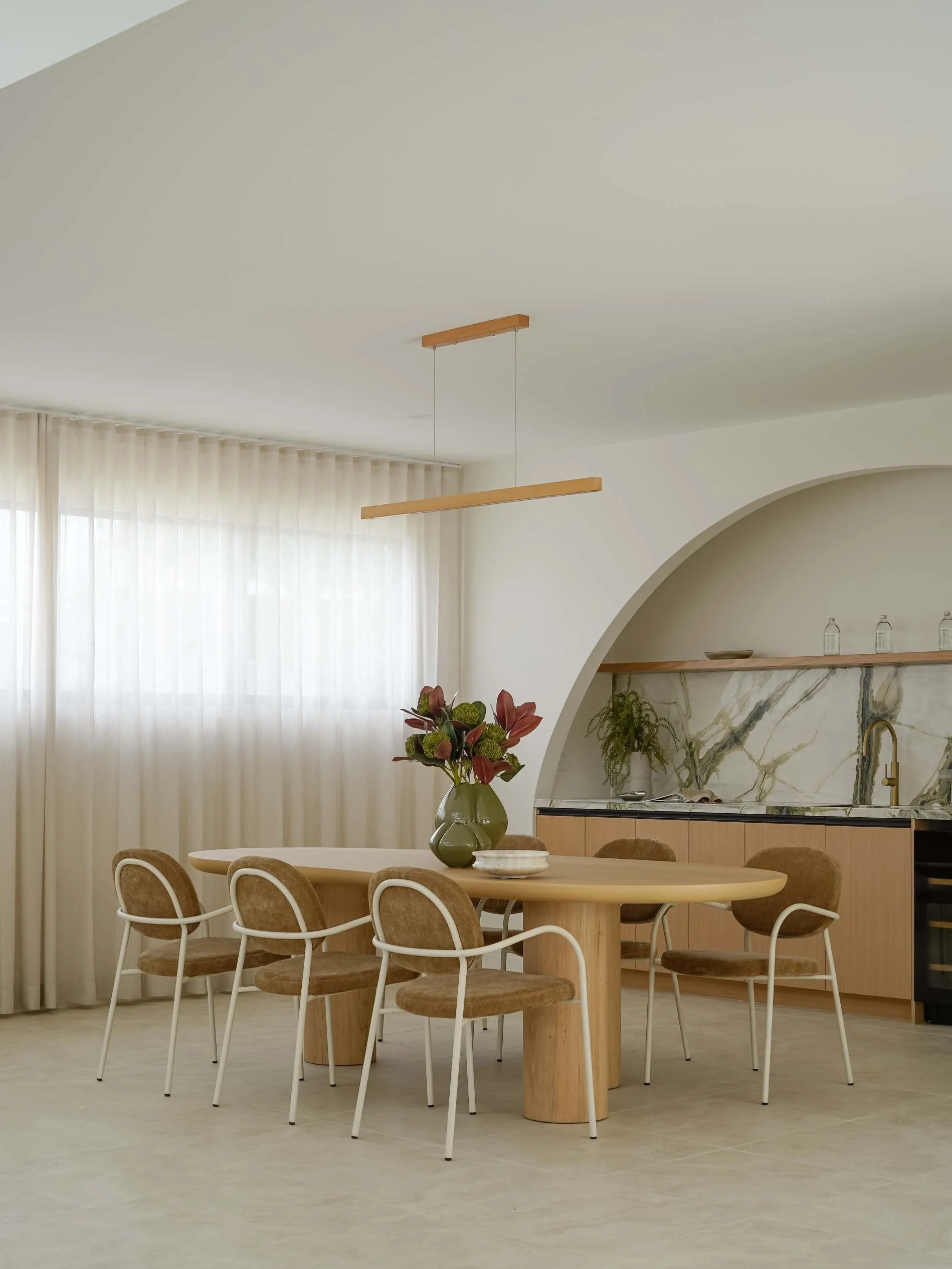 Modern dining room with a round wooden table, six beige chairs with white metal frames, a vase with large pink and green flowers, large windows with sheer white curtains, a curved wall with a wooden shelf and marble backsplash, and minimalistic decor