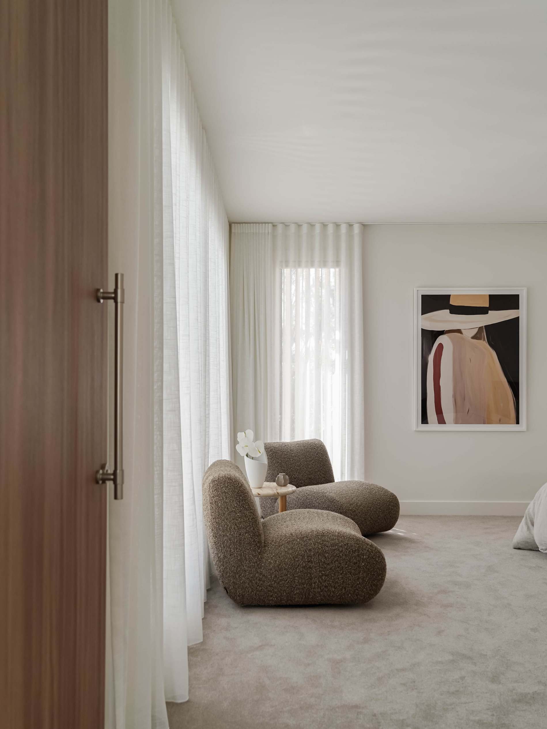 A minimalist living room corner with two brown upholstered chairs, a small round side table, a white vase with a white orchid, a painting of a woman wearing a large hat on the wall, and sheer white curtains covering large windows.