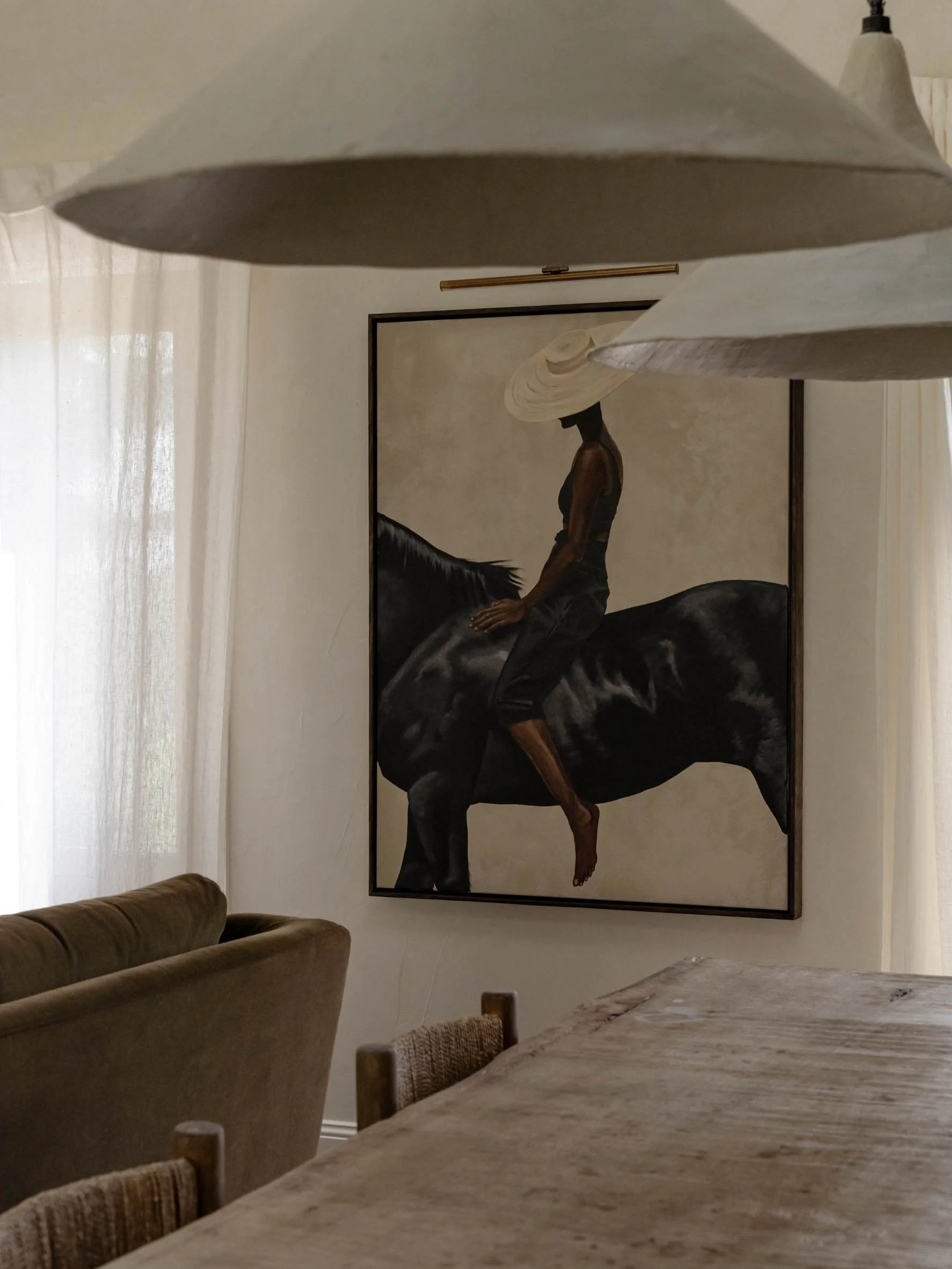 A framed artwork of a black horse with a woman riding it, positioned on a light-colored wall in a room with natural light, a dining table, and hanging lamps.