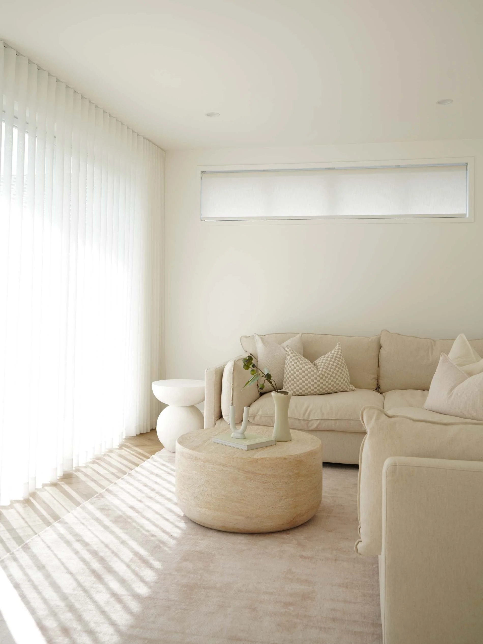 Bright living room with white walls, a beige sectional sofa with patterned and plain cushions, a round beige coffee table with a white book and vases, sheer white curtains, and minimal decor in Ostani House Gold Coast