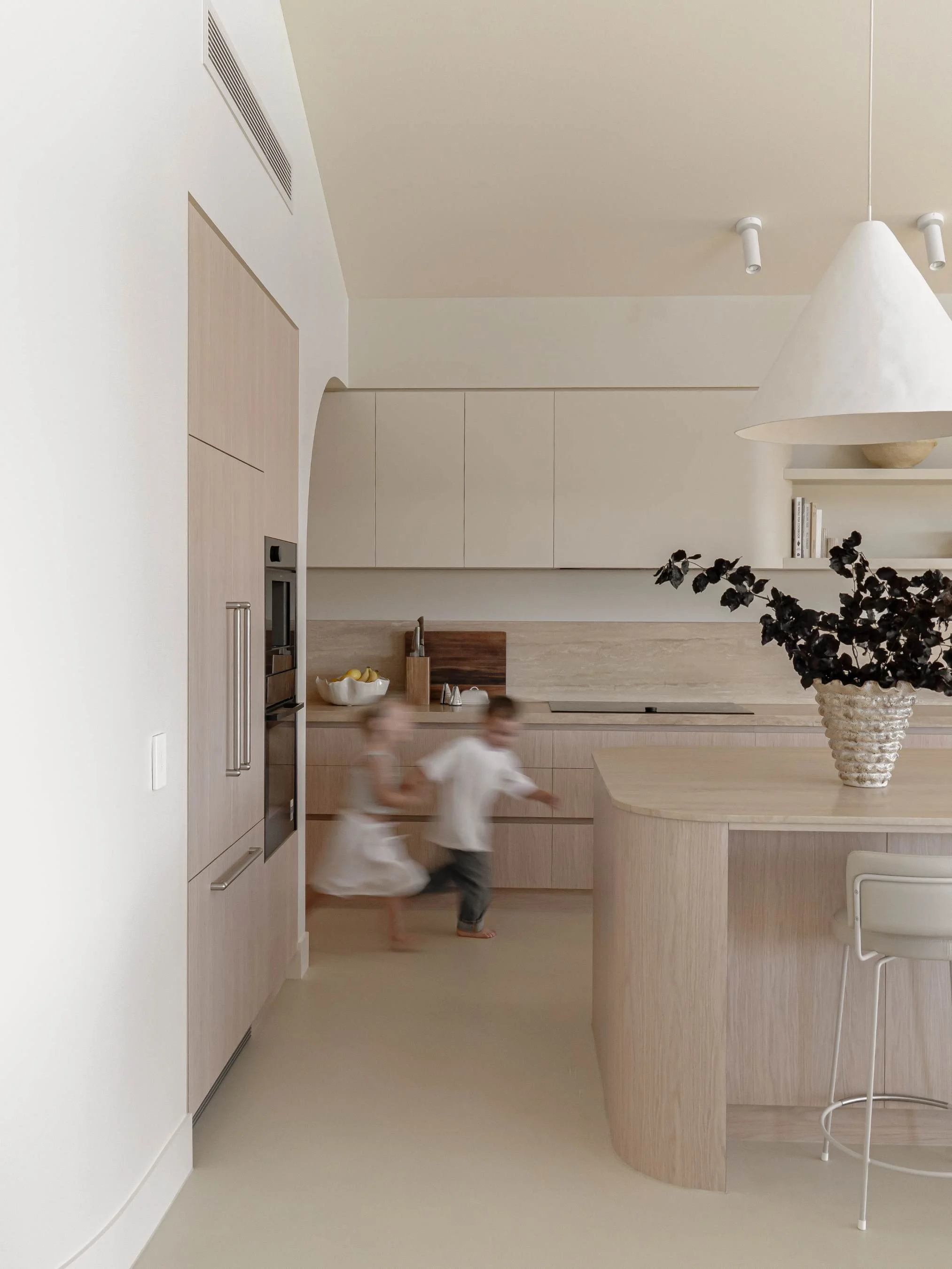 Two children running in a modern, minimalist kitchen with light wood cabinets and neutral tones in the Ocean Ave Project in Tweed Heads, QLD
