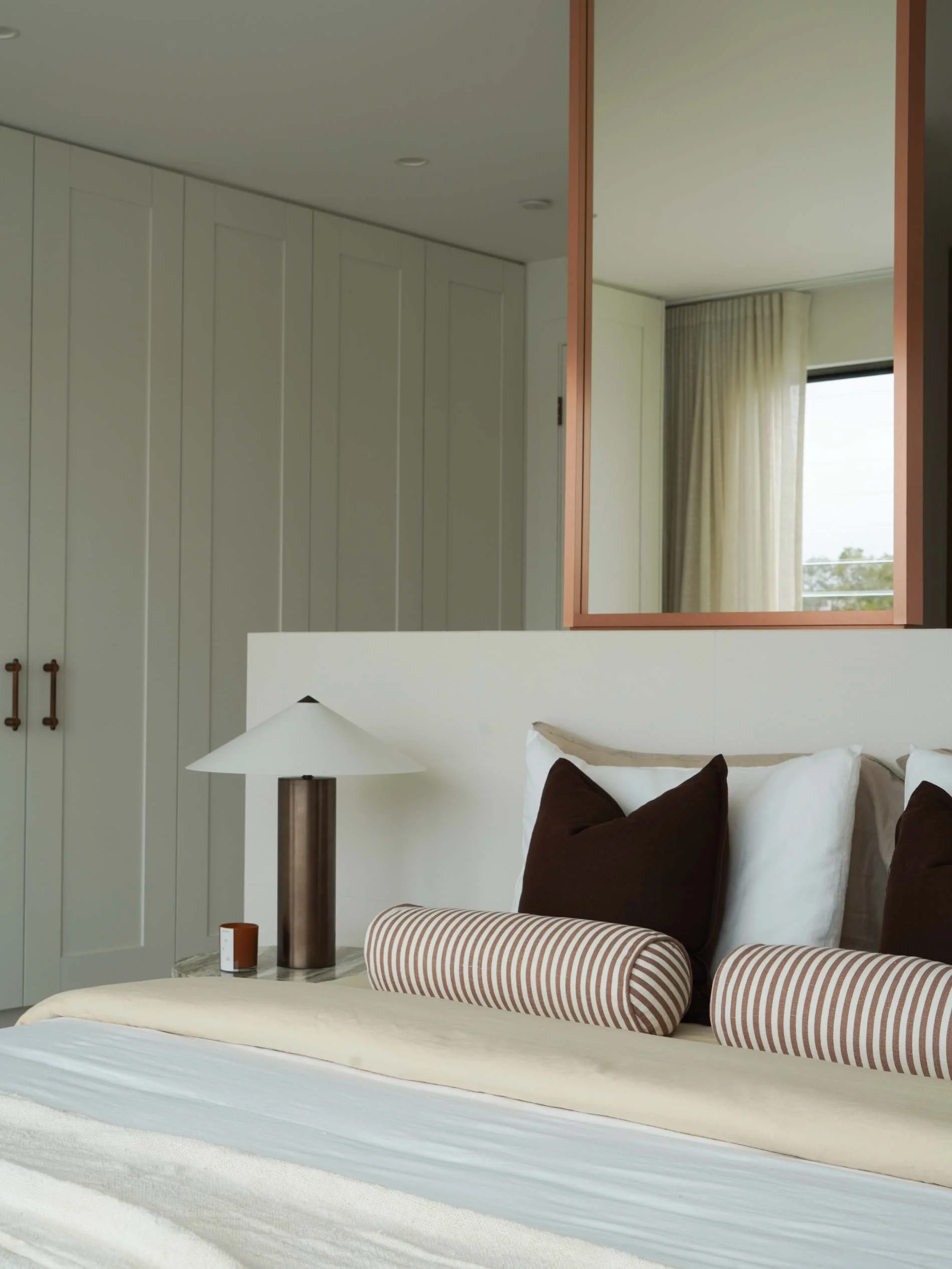A mid century bedroom with a bed, dark brown and white pillows, a modern lamp, a large mirror, a window with curtains, and built-in closet cabinets in honeyeater drive burleigh waters