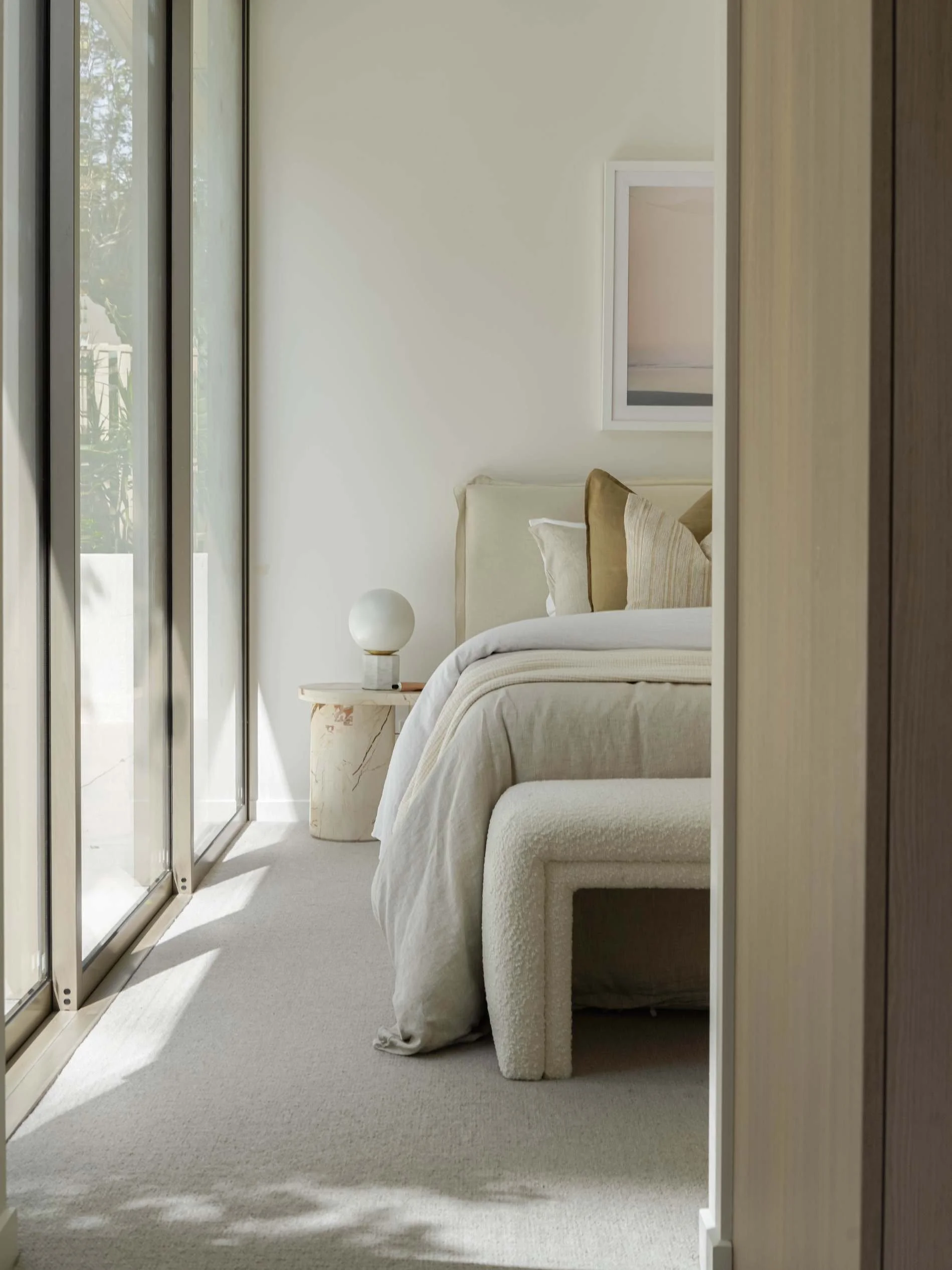 A modern bedroom with a bed, beige and gold pillows, a white blanket, a round marble nightstand with a white spherical lamp, near large sliding glass doors and a minimal framed artwork on the wall at Florence, Mosaic Developers in Burleigh Heads