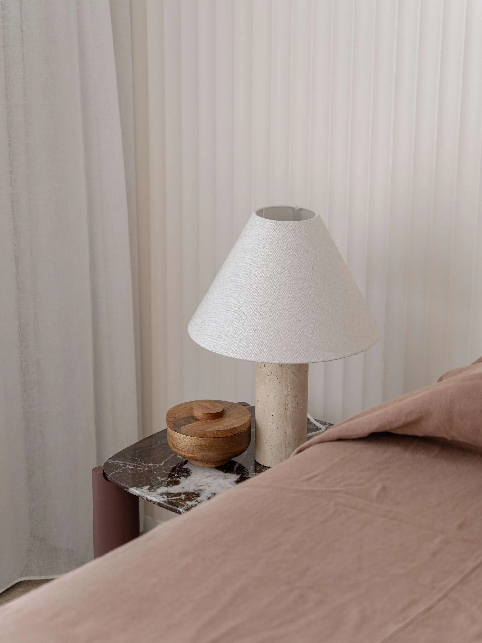 Nightstand with a wooden container and a table lamp next to a bed with a pink bedsheet and sheer white curtains in the background.