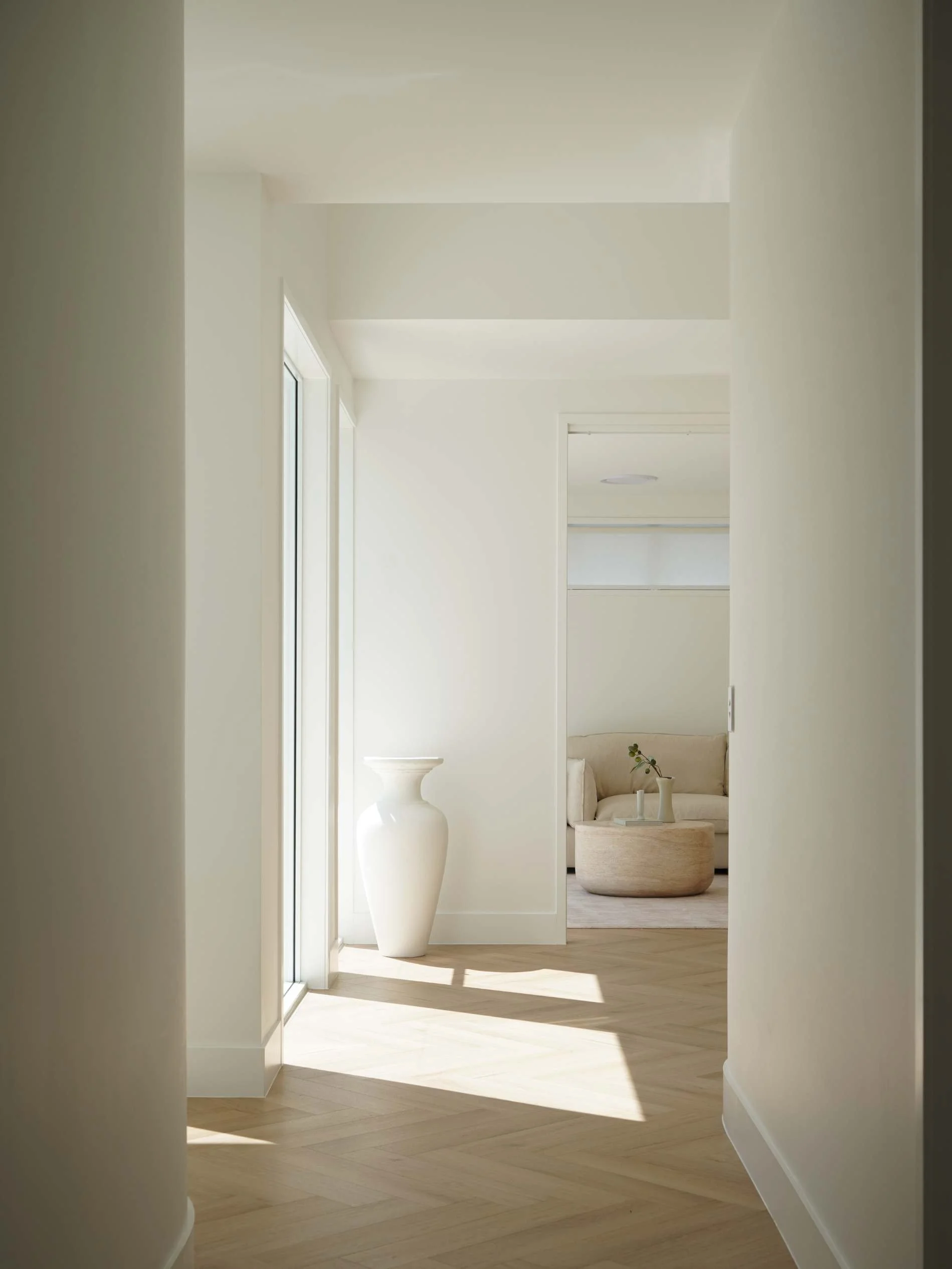 A bright, minimalistic hallway with light-colored wooden flooring, white walls, and a large white vase near a glass door. In the background, a cozy seating area with a sofa and a round ottoman, decorated with a vase and a plant, is visible.