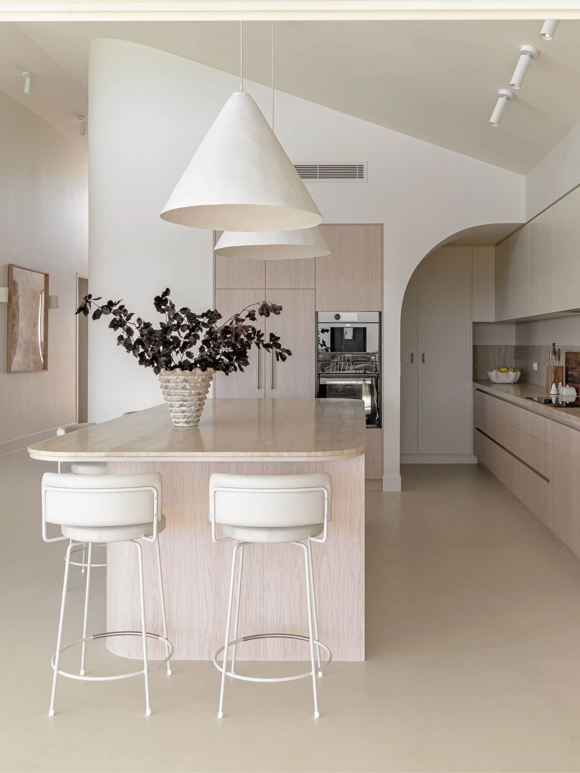 Modern minimalist kitchen with a central island, white bar stools, and a large white pendant light above. A vase with dark foliage decorates the island, with built-in appliances and simple cabinetry in the background in the Ocean Ave Project in Tweed