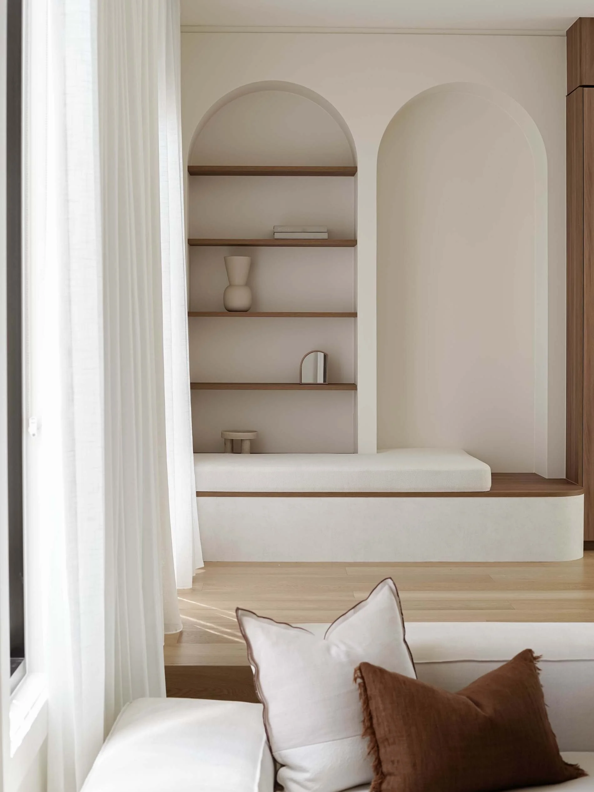 Living room with built-in white sofa with white and brown pillows, wooden floor, and white curtains. A built-in shelving unit with decorative items and a mirror is visible in the background in Savoy, Broadbeach Waters, QLD 