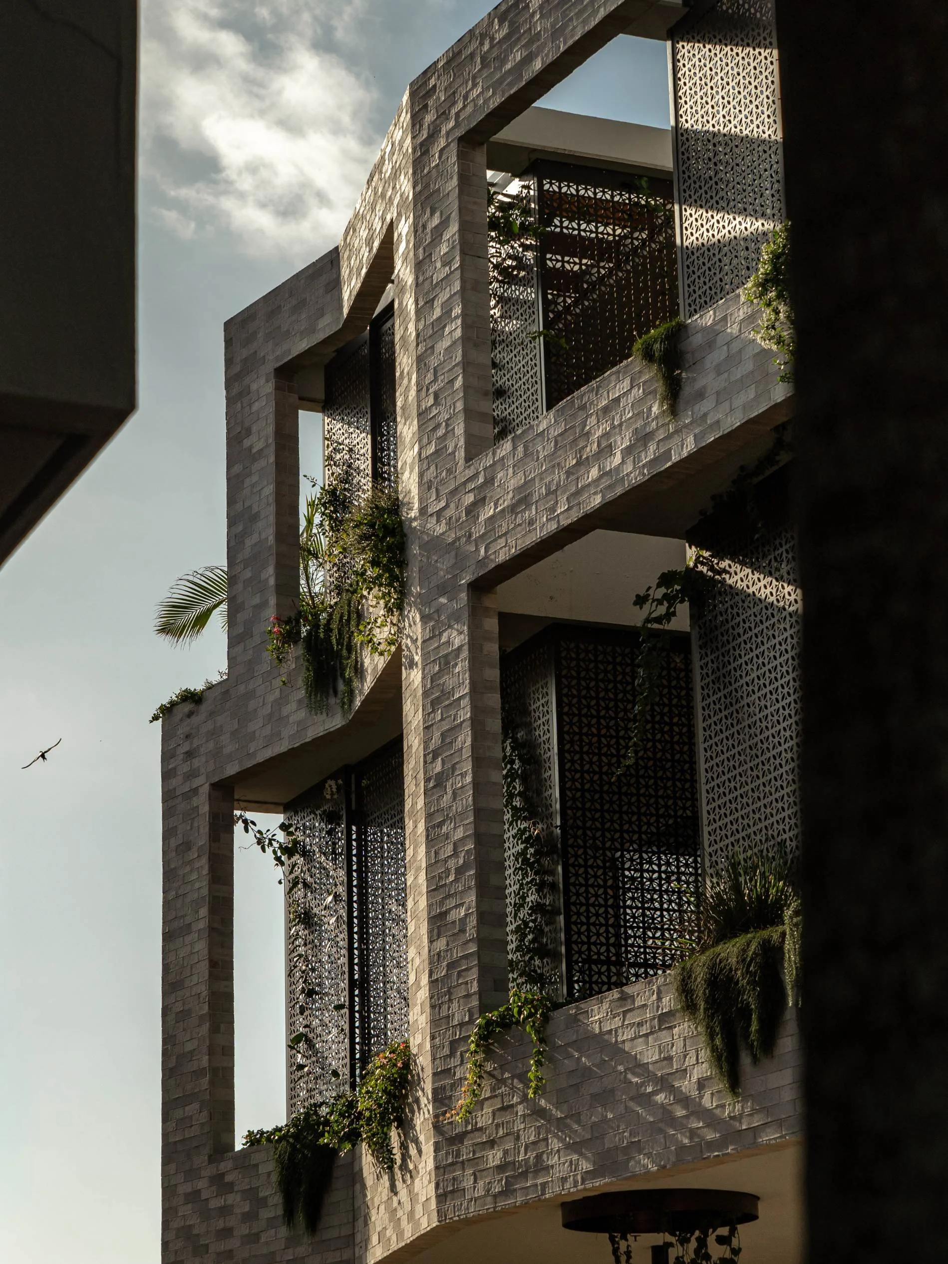 A modern multi-story building with a facade made of light gray bricks, featuring large open balcony spaces with decorative black metal screens, green plants, and foliage on the balconies. The sky is partly cloudy with an airplane flying in the distan