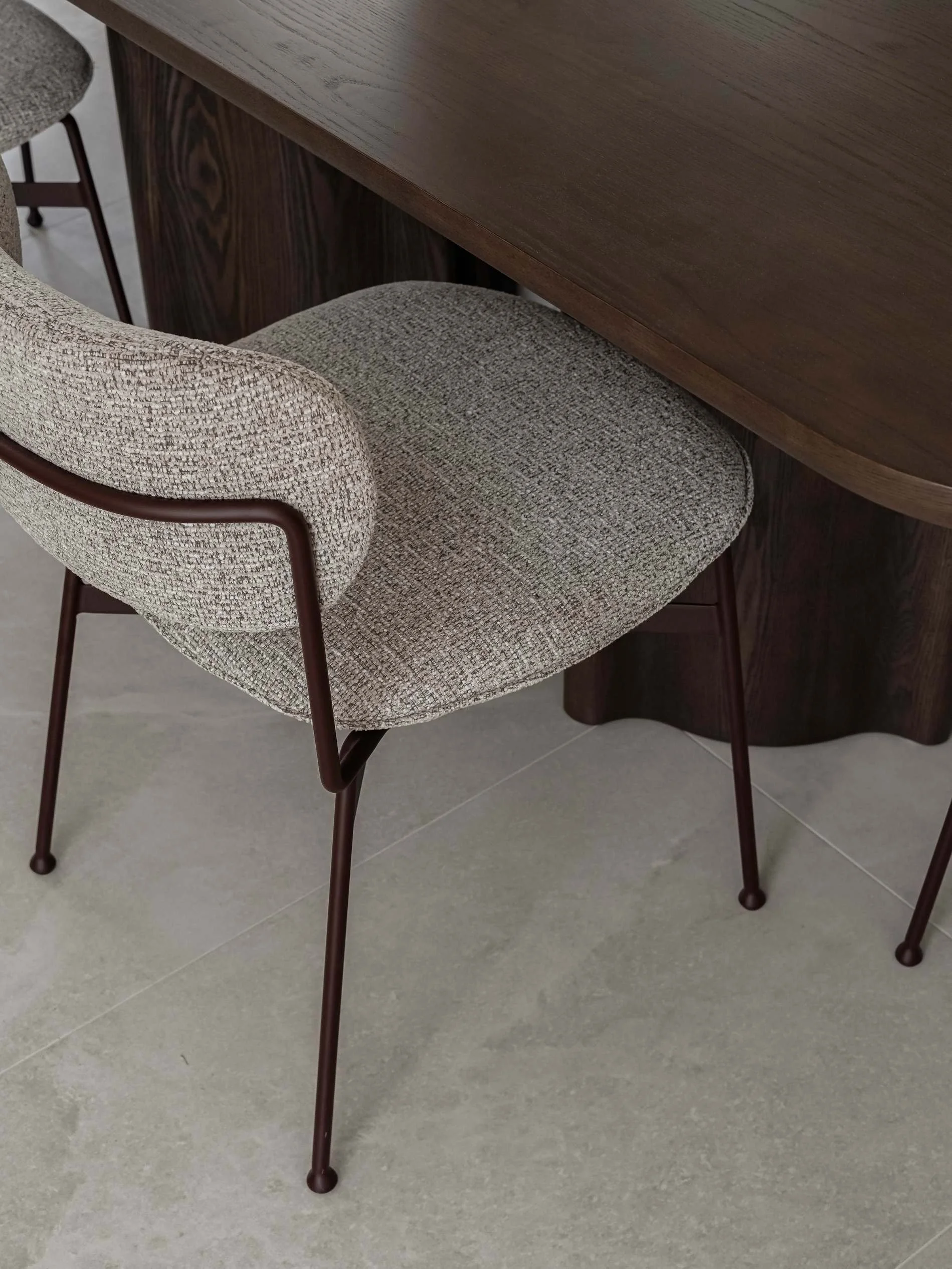 Close-up of a modern dining chair with beige textured fabric upholstery and dark metal legs, partially tucked under a wooden table with a dark finish in Savoy, Broadbeach Waters, QLD 