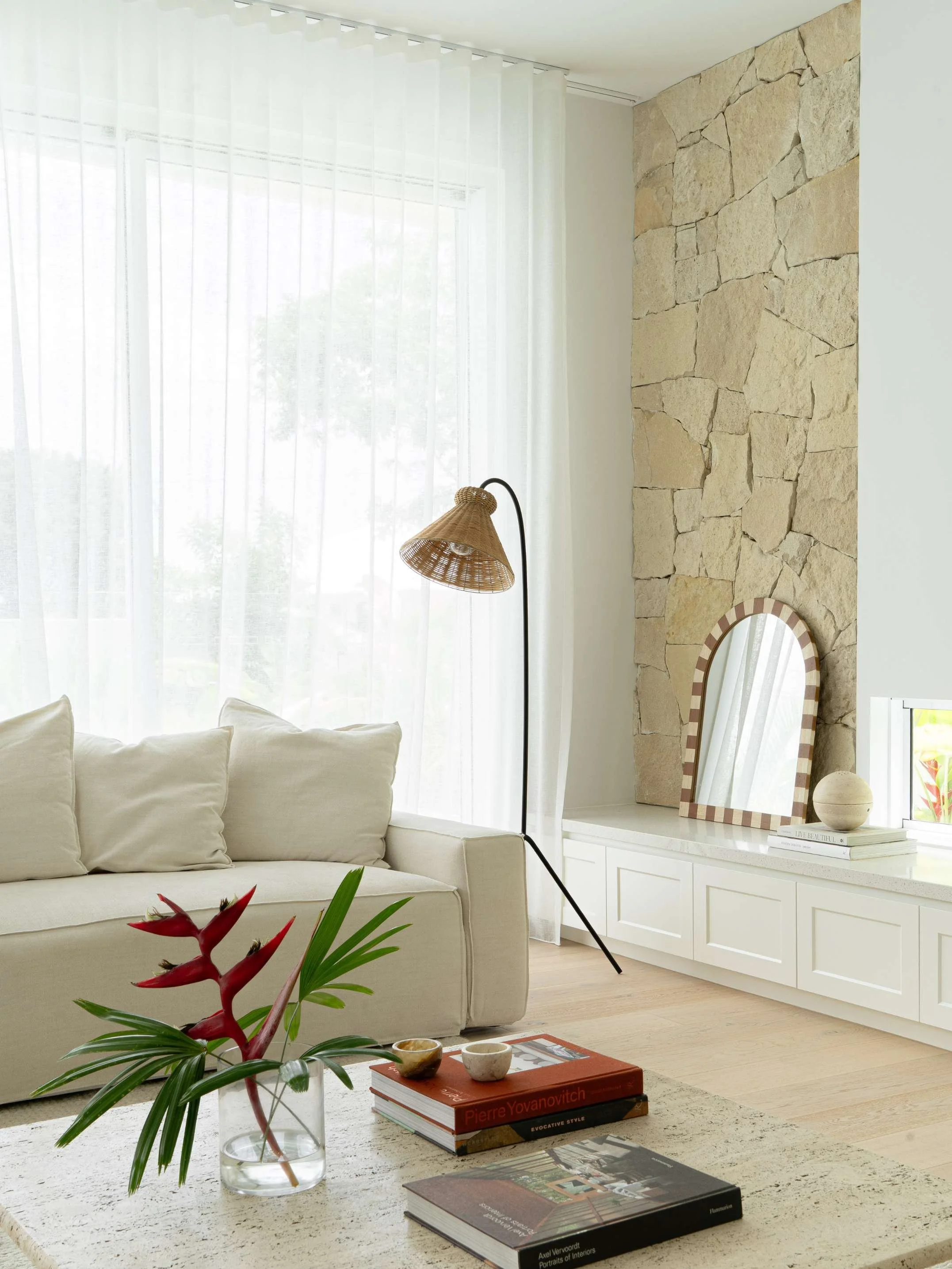 Bright living room with a white sofa, floor lamp, stone accent wall, decorative mirror, and books with small bowls on a coffee table.
