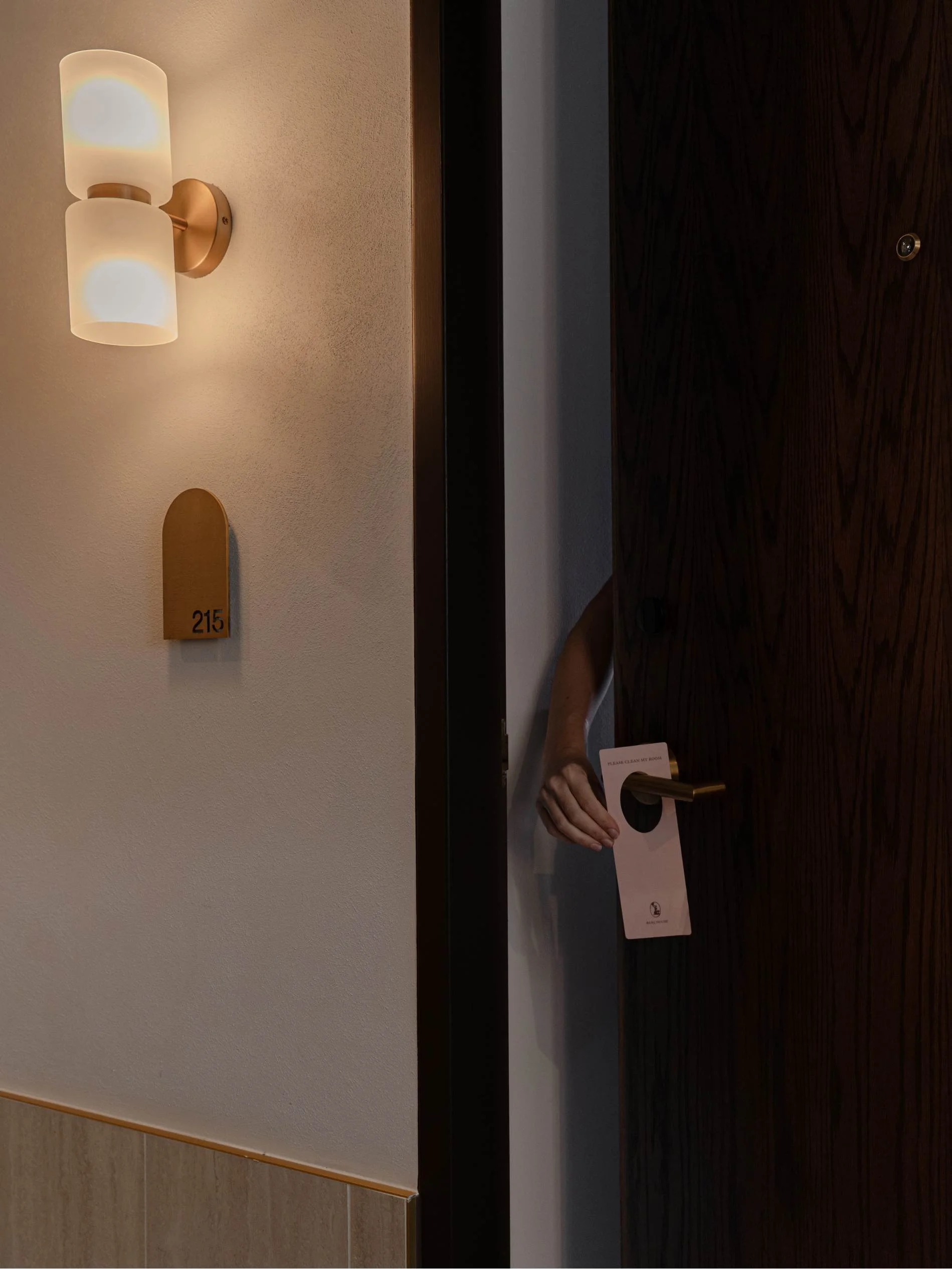 Partially opened hotel room door with a hand holding a door hanger that says 'Please Clean My Room.' The door has a peephole. To the left, a wall-mounted light fixture with two frosted glass shades and a room number plaque '215'.