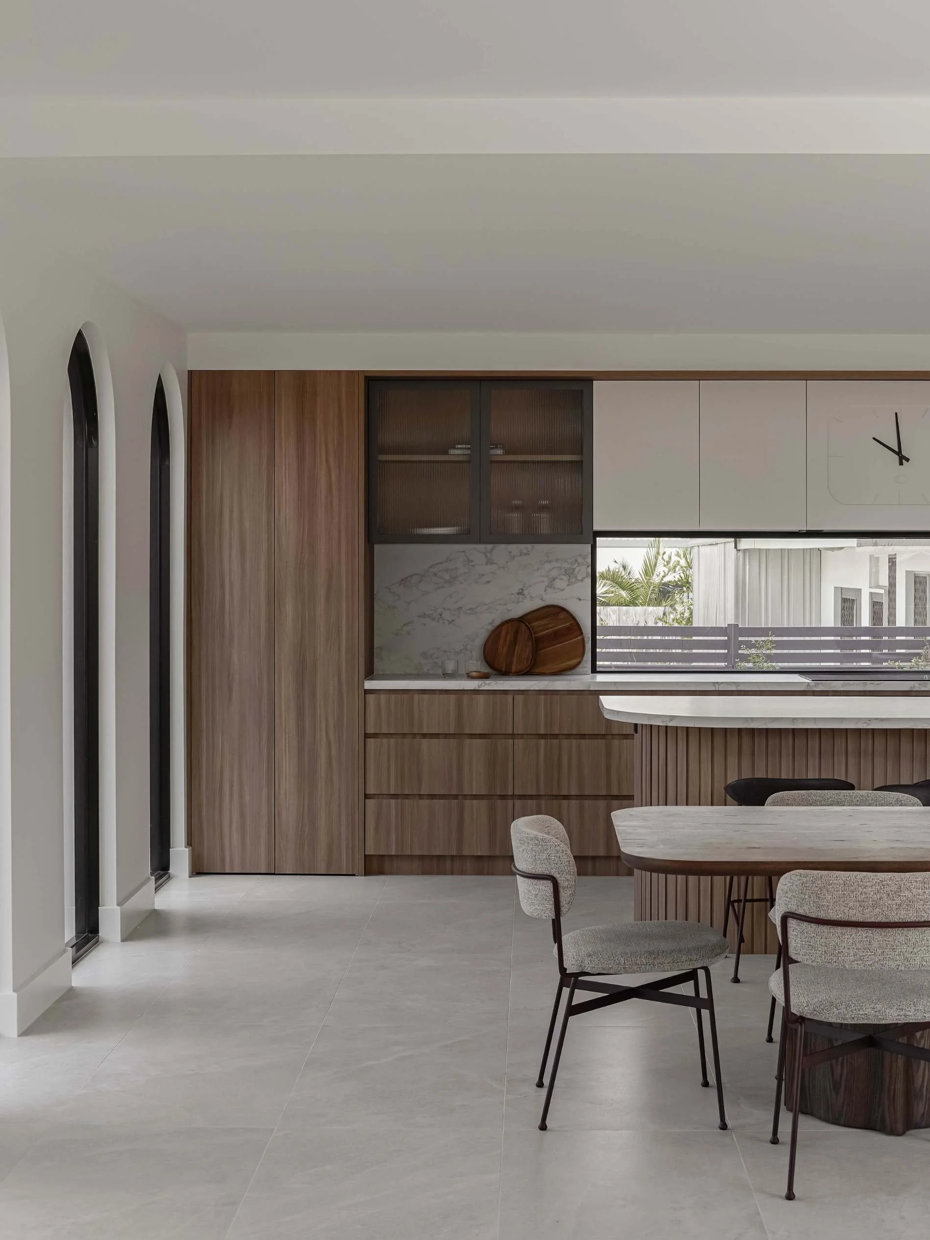 Modern kitchen with wooden cabinets, marble backsplash, and a window overlooking outdoor greenery, with a dining table and chairs in the foreground in Savoy, Broadbeach Waters, QLD 